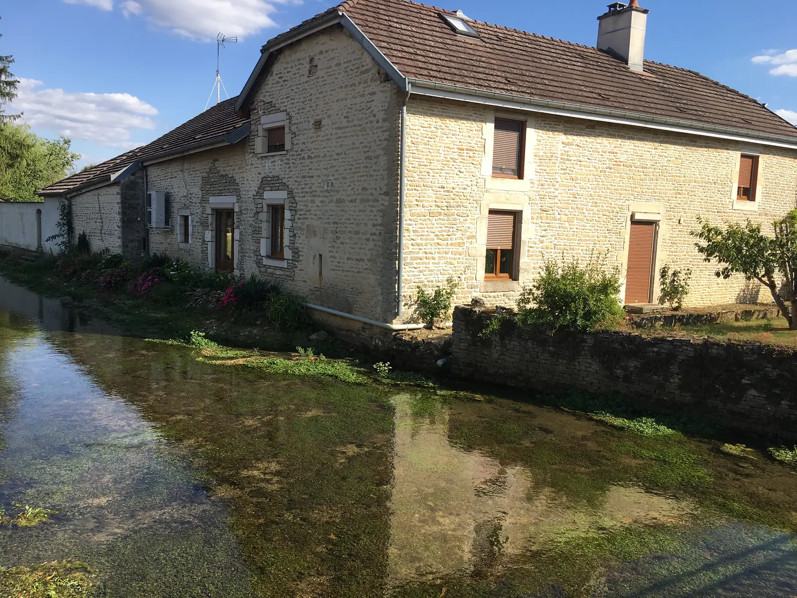 A stone farmhouse with shuttered windows and flowering shrubs sits beside a clear, weed-filled stream running through the village.