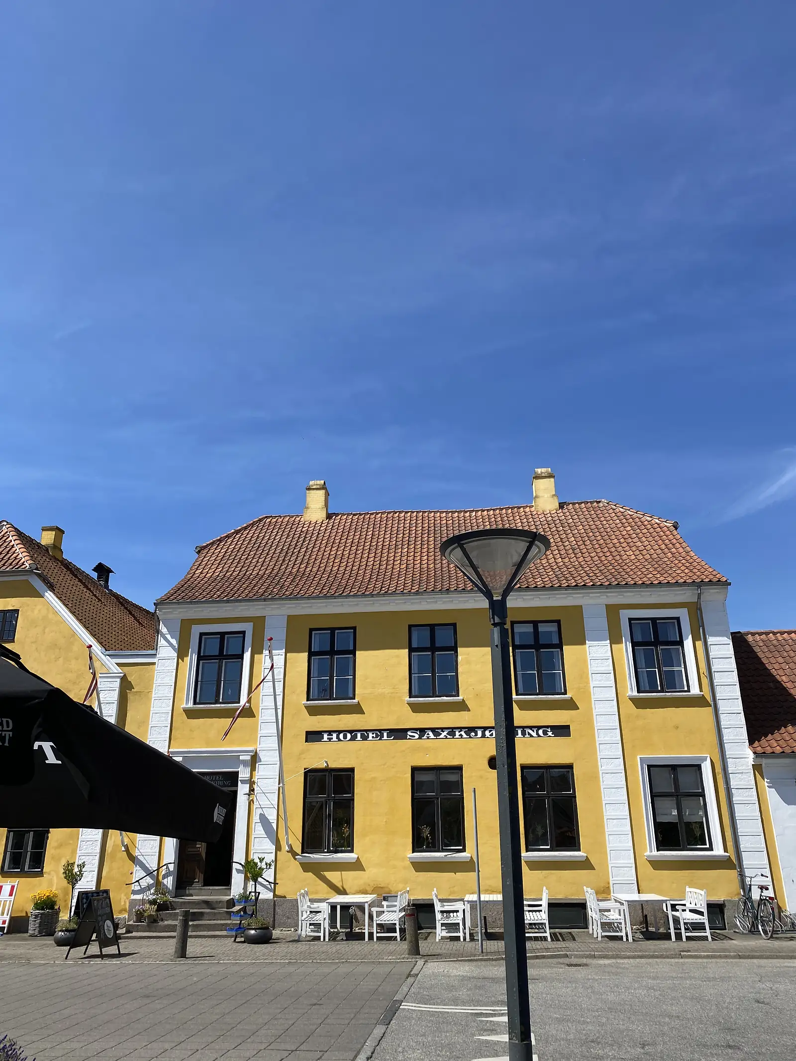 A yellow rendered hotel with a red-tiled roof lettered "HOTEL SAXKJØBING", white cafe chairs outside under a deep blue sky.