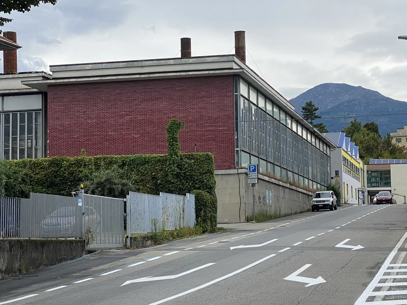 A red-brick industrial building with tall chimneys and ivy climbing its wall, glazed workshop windows running along its side towards distant mountains.