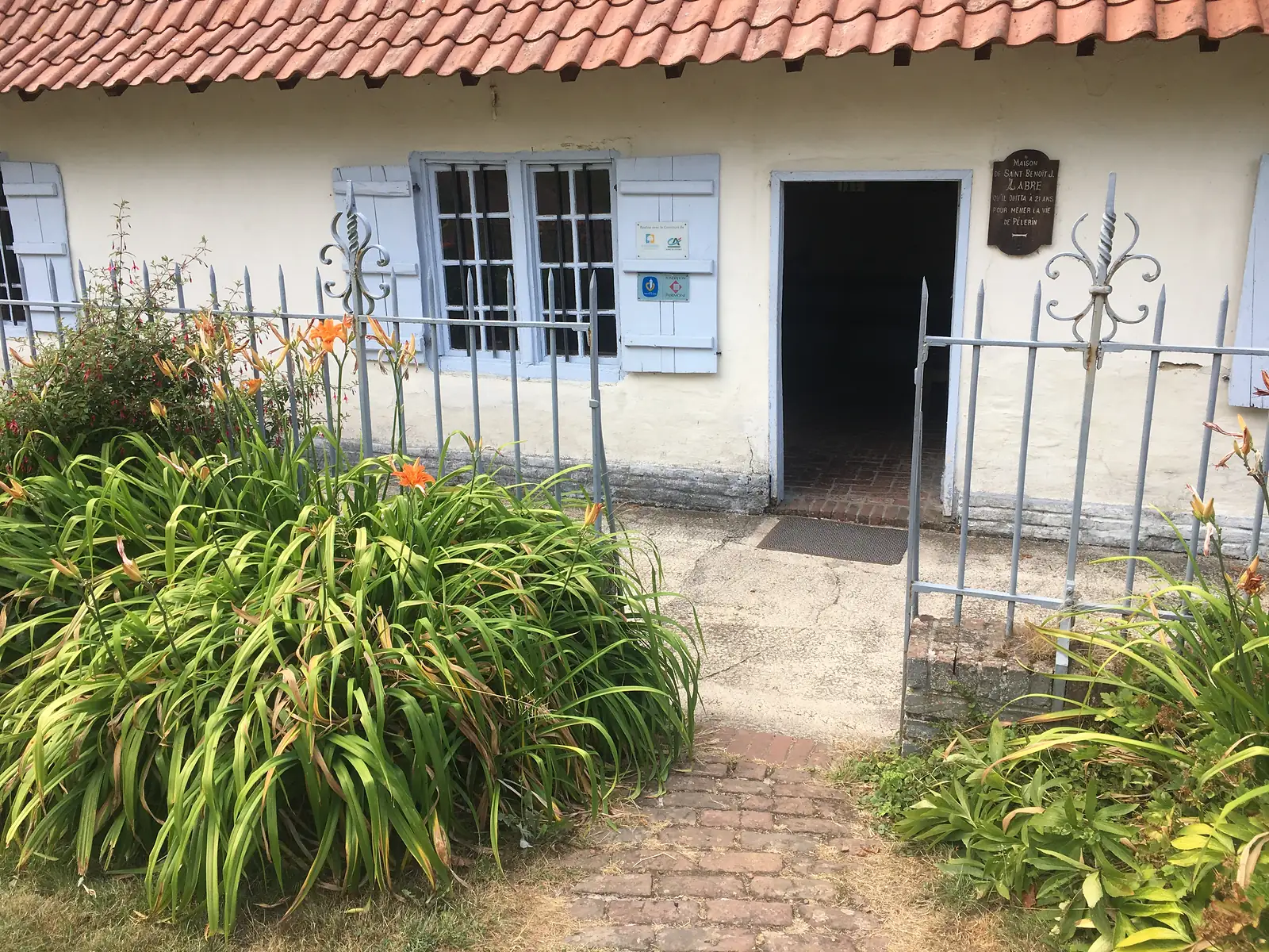 The whitewashed, blue-shuttered cottage where Saint Benoît Joseph Labre was born, its open doorway framed by a plaque reading 'Maison de Saint Benoît J. Labre'.