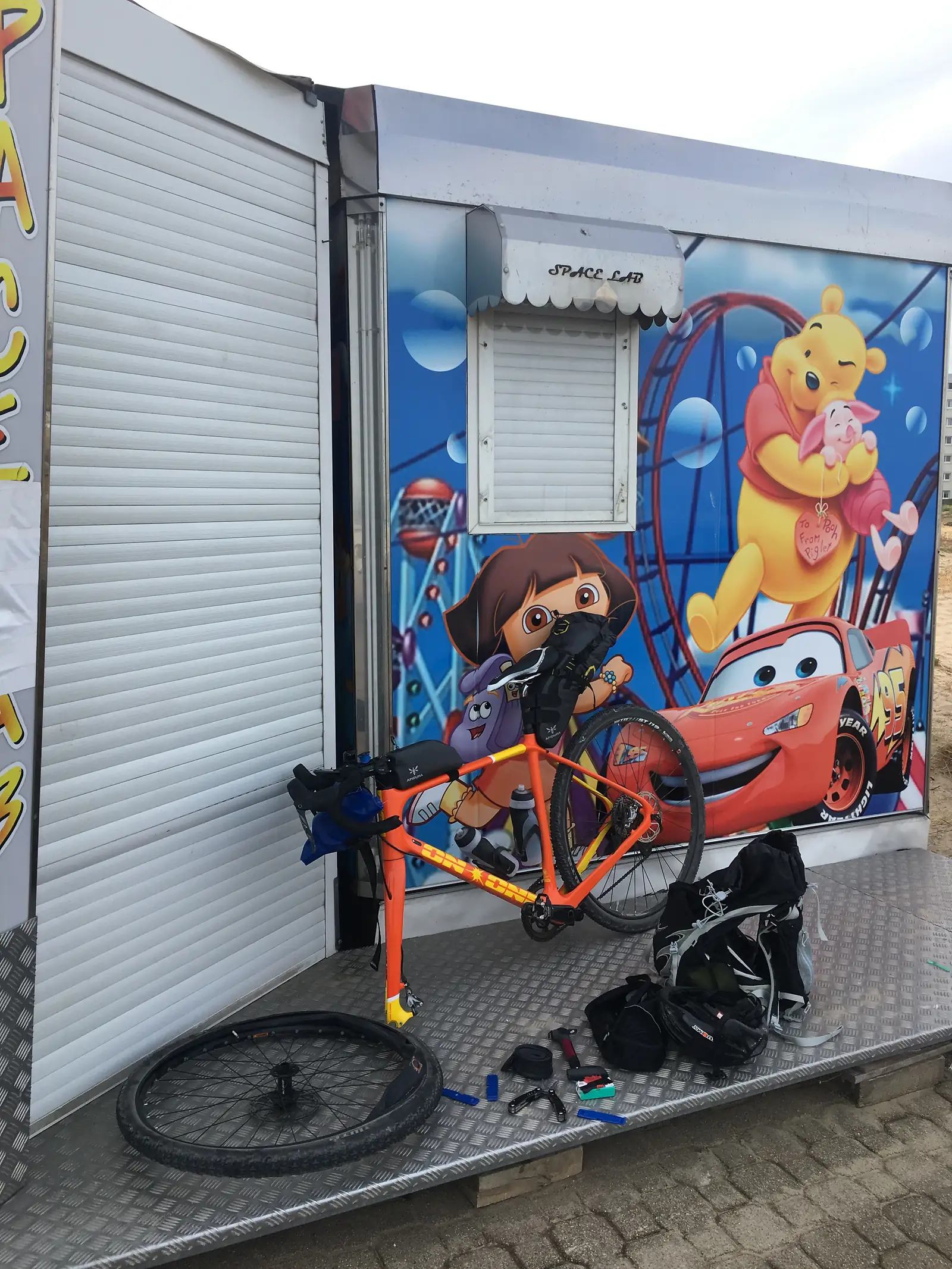 An orange bike propped against a shuttered shopfront painted with cartoon characters including Winnie the Pooh and Lightning McQueen, panniers and gear stacked alongside on a chequer-plate ramp.