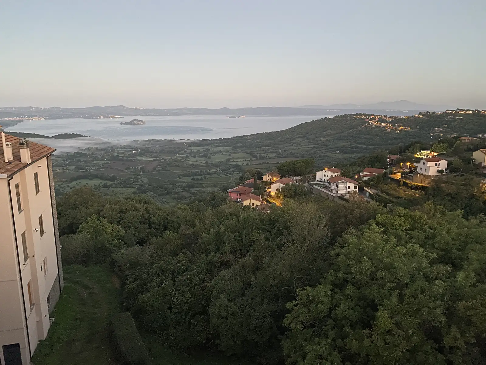 Dawn or dusk view over a large lake from a hillside town, with red-roofed houses among trees in the foreground and mist settling over fields near the shore.