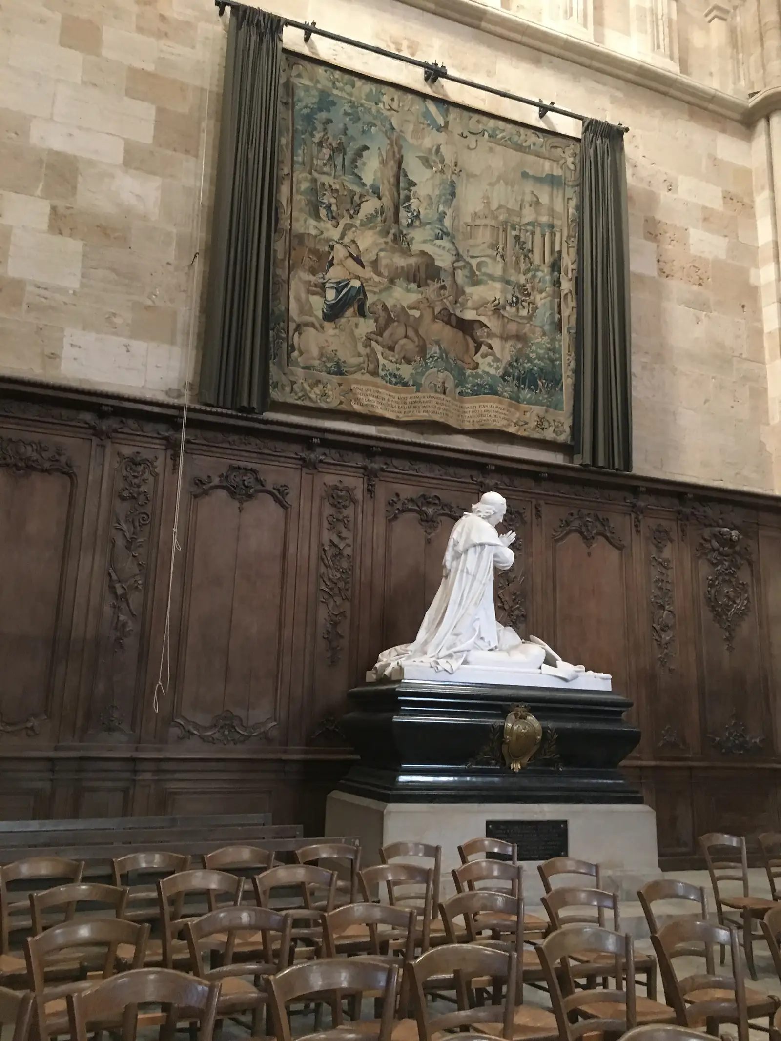 A white marble bust on a dark plinth beside a faded tapestry and carved wood panelling, with rows of chairs in the foreground of a church interior.