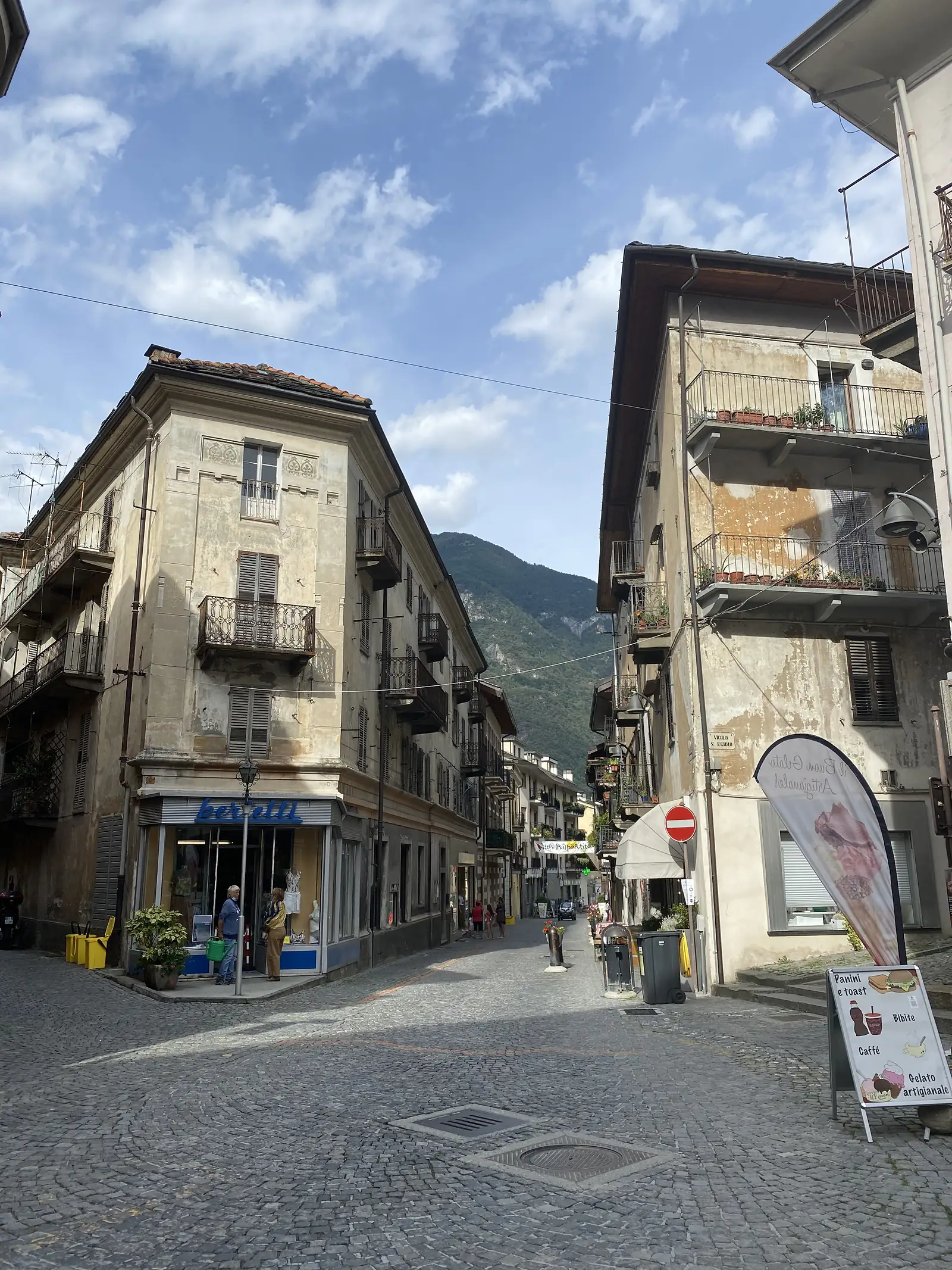 A cobbled Italian street corner with weathered ochre and cream buildings, shuttered windows, and a couple standing outside a shopfront beneath a mountain backdrop.