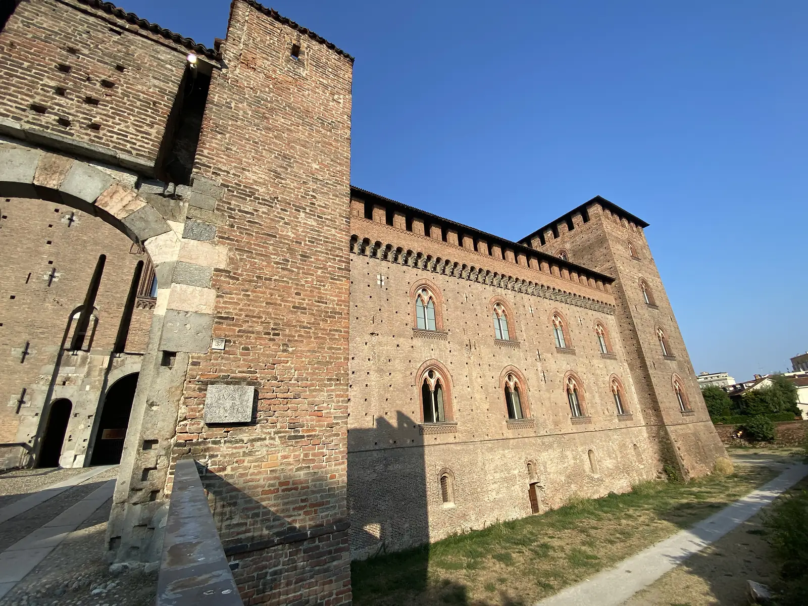 The crenellated brick tower and walls of a medieval castle rise above a gravel path, a plaque reading 'MUSEO CIVICO' fixed to the corner tower.
