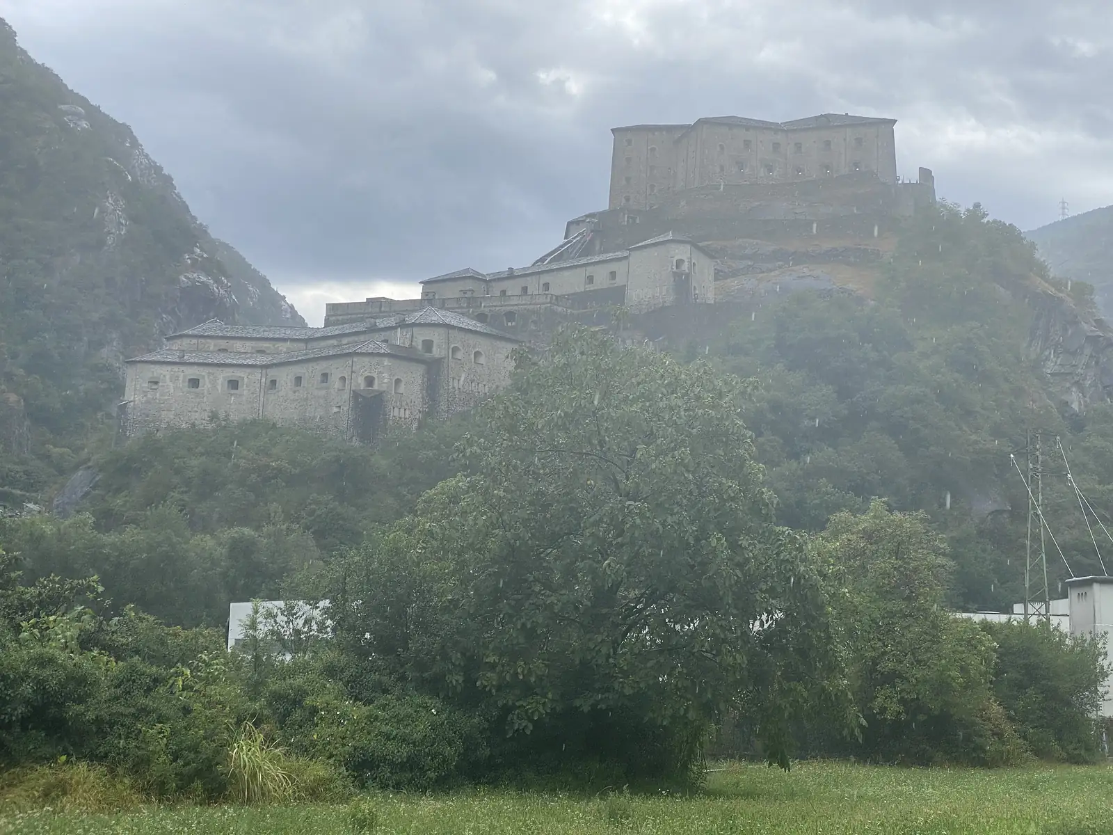 The stacked stone bastions of the Fort of Bard rising above woodland on a misty hillside, photographed through light rain.