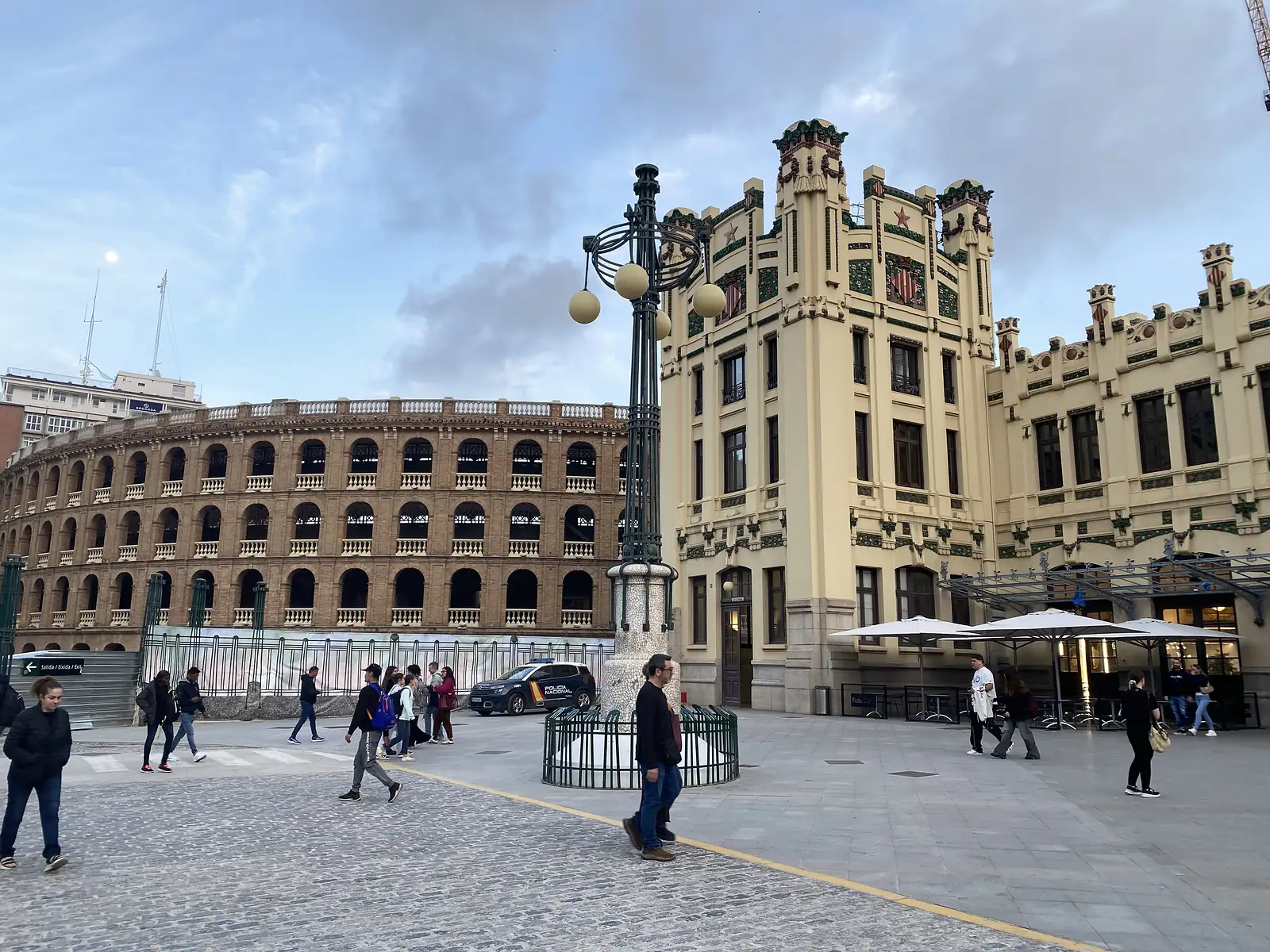 A brick-arcaded bullring standing beside an ornate art nouveau railway station, people crossing the paved square between them.