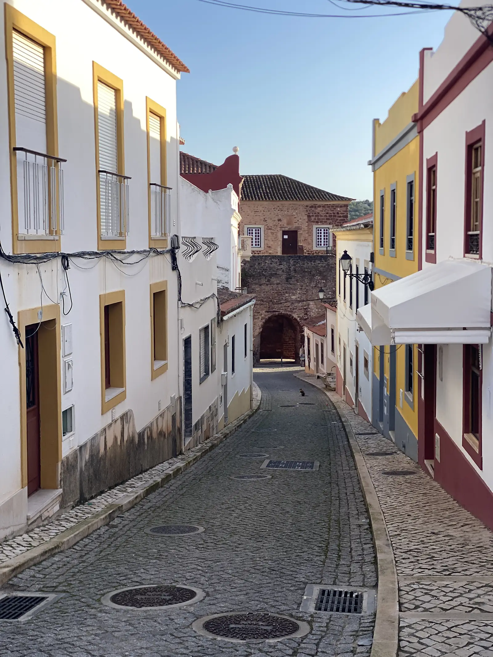 Cobbled street of yellow- and red-trimmed houses sloping down to an arched gateway in a red sandstone castle tower at the end.