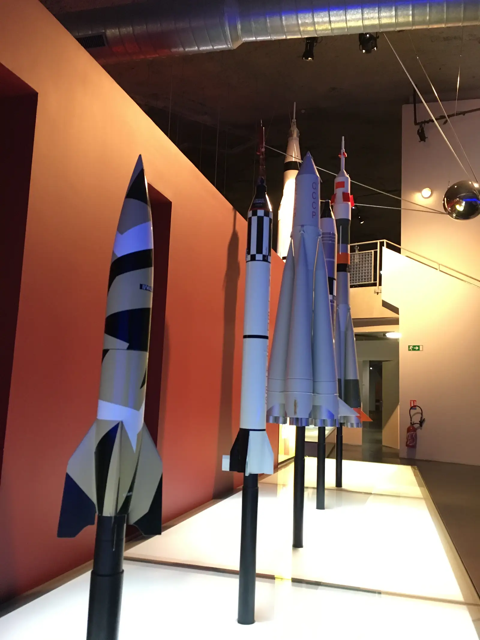 A row of small rocket models, one lettered 'СССР', stand nose-up along a museum display stand beneath spotlights.