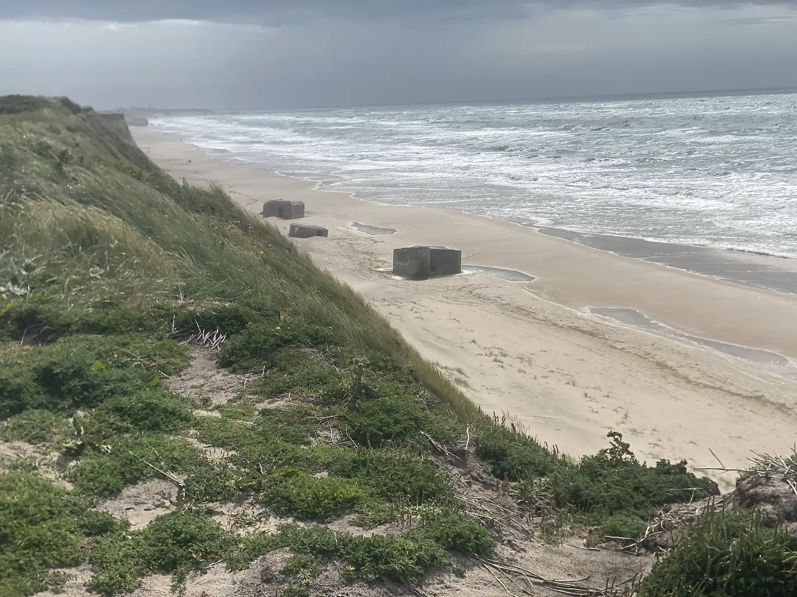 Concrete bunkers slumped onto the sand below an eroding grassy dune cliff, with a rough grey sea breaking along the shore.
