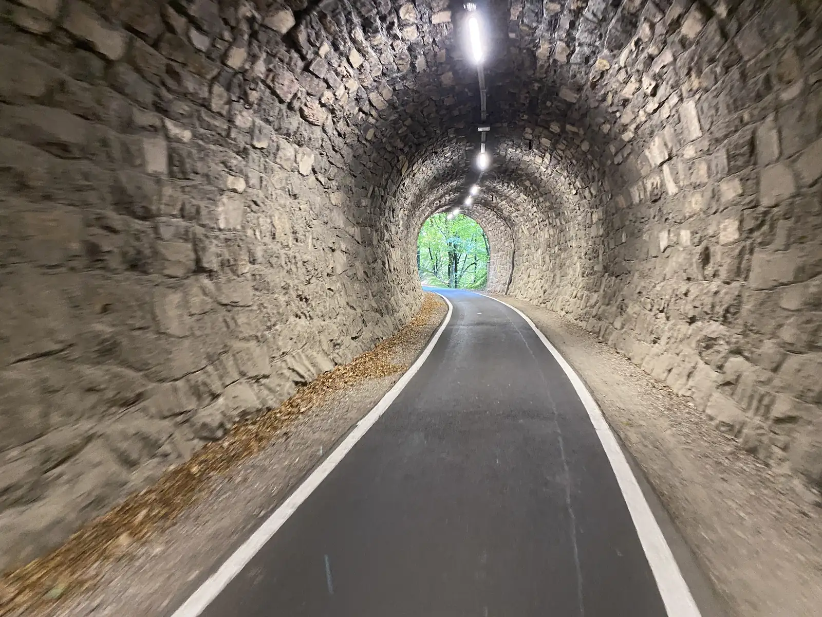 A lit, stone-lined tunnel carries a tarmac cycle path, its white edge lines curving towards green daylight at the far portal.