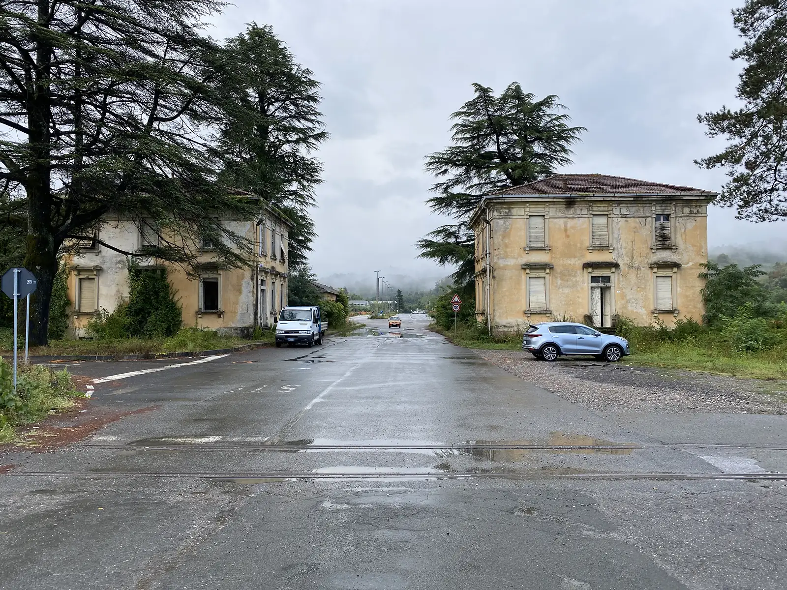 Two derelict ochre villas with shuttered windows and tall cedars flank a wet crossroads, a van and a car parked outside as mist fills the road ahead.