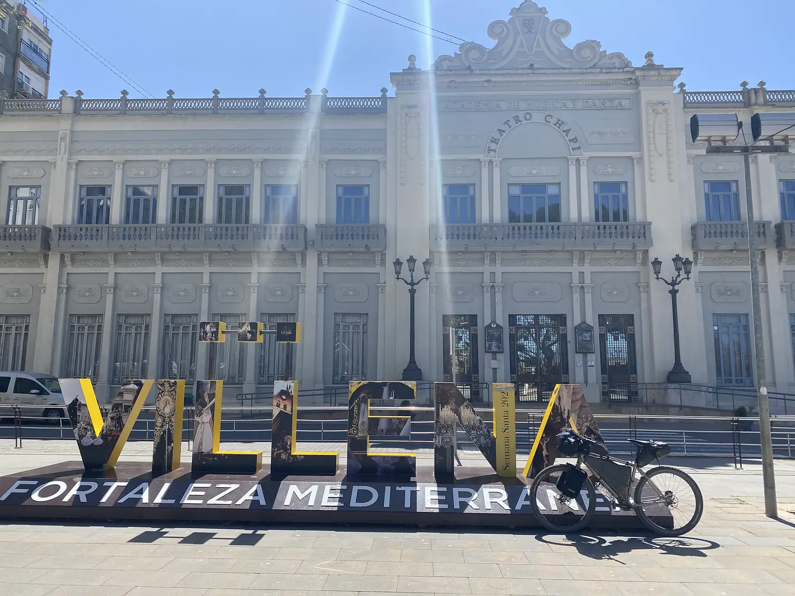 A loaded bike propped against giant letters spelling "VILLENA" on a plinth reading "FORTALEZA MEDITERRANEA", in front of a grand white theatre lettered "TEATRO CHAPI".