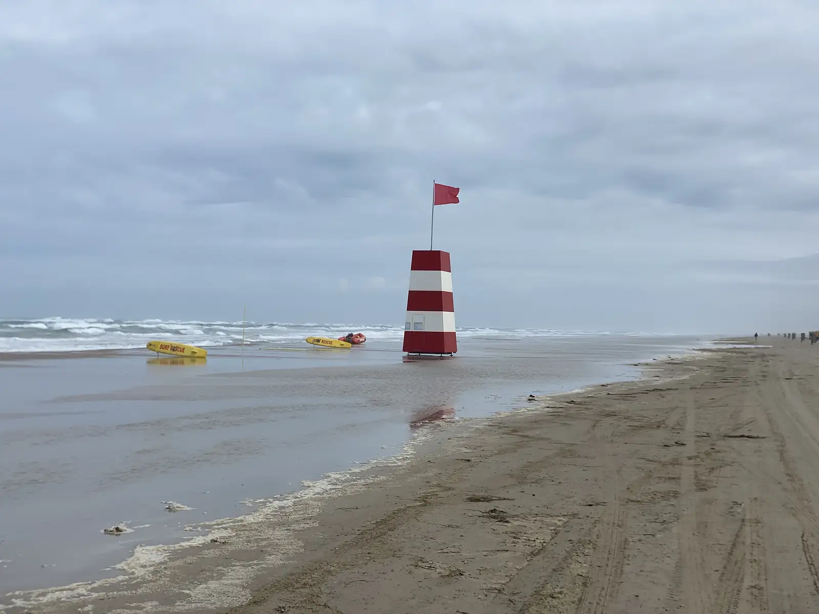 A red-and-white striped lifeguard tower flying a red flag on a wet beach, with yellow rescue boards lettered "SURF RESCUE" on the sand.