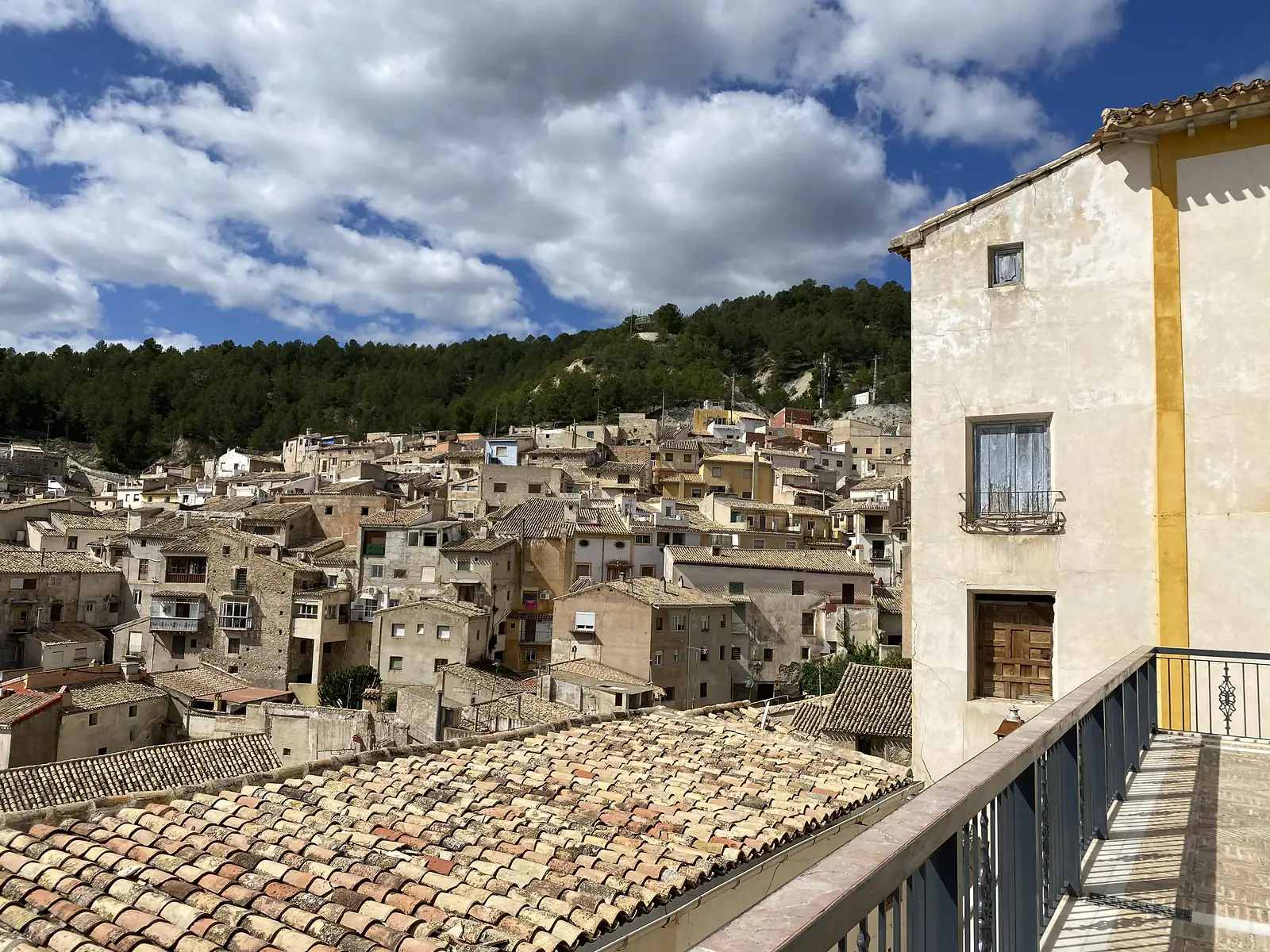 Tiled rooftops of a town packed up a pine-covered hillside, seen from a balcony beside a cream and yellow church wall.