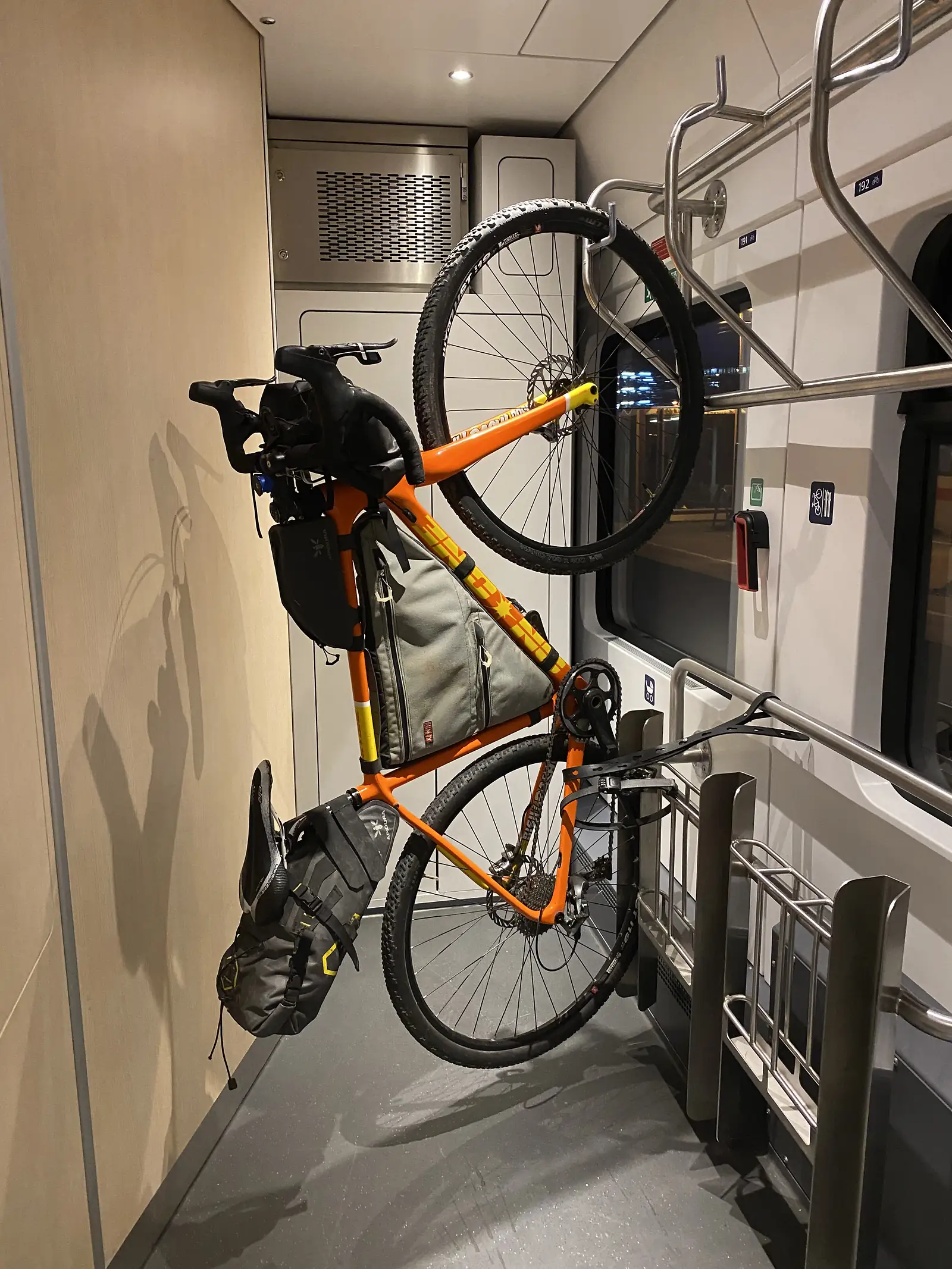 A loaded orange gravel bike hangs from a hook in a train's designated bicycle bay, marked by a bike symbol on the wall.