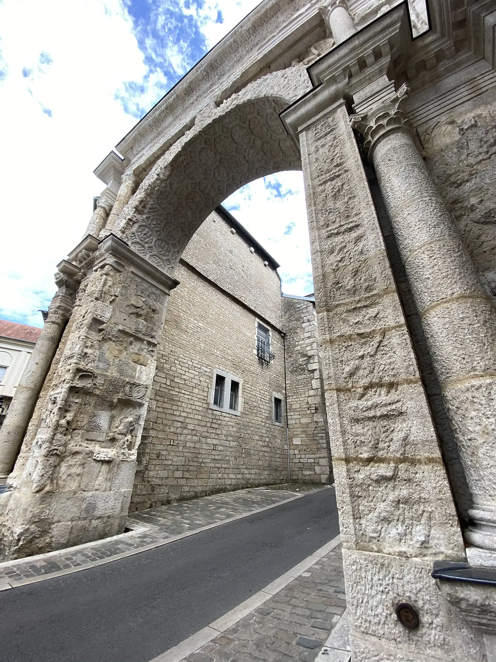 A weathered stone Roman triumphal arch with ornately carved surfaces frames a view of a stone building and quiet street below.