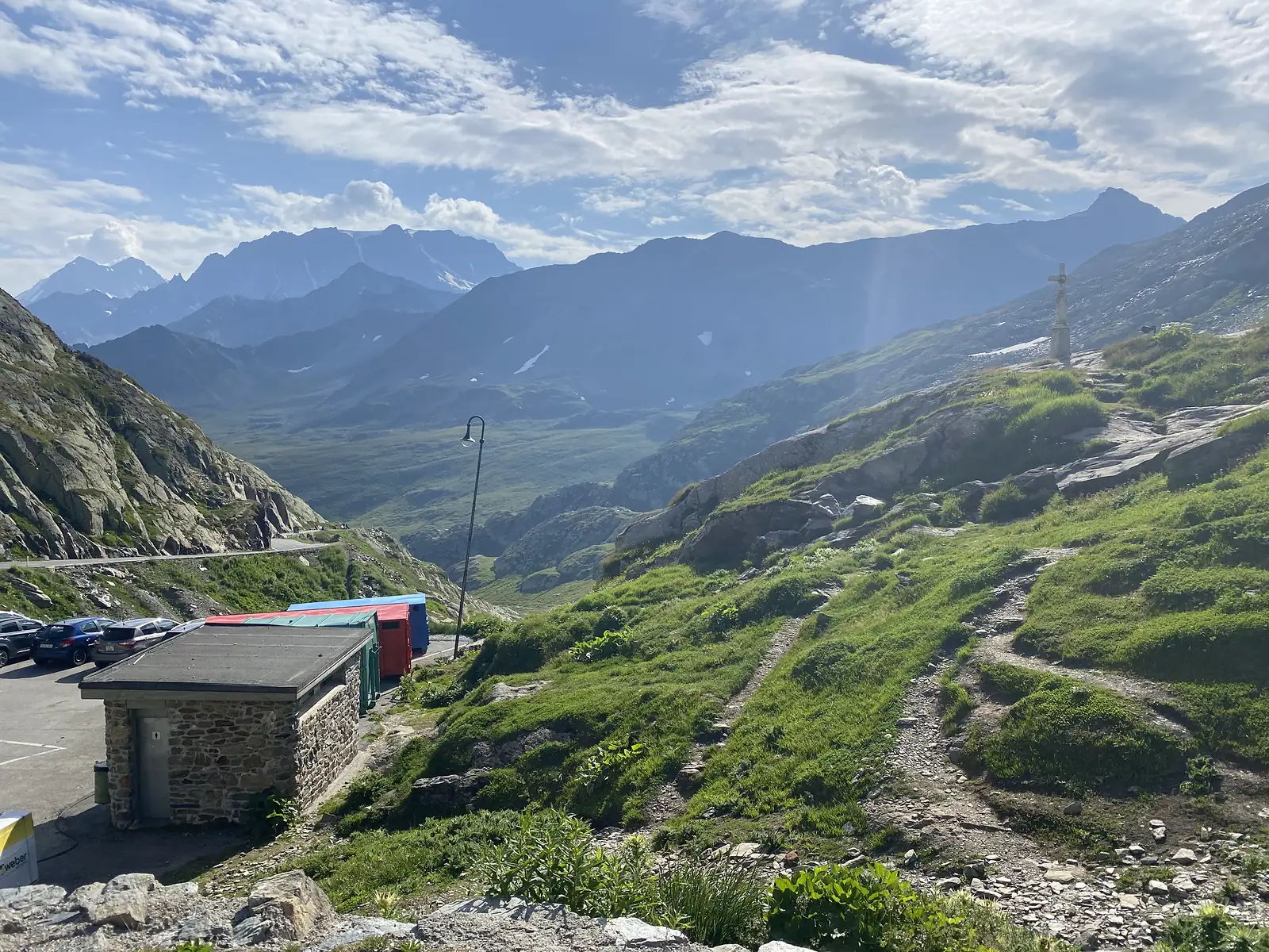 A stone toilet block and colourful storage sheds beside a mountain-pass car park, with a rocky trail and a stone cross climbing the grassy slope beyond.