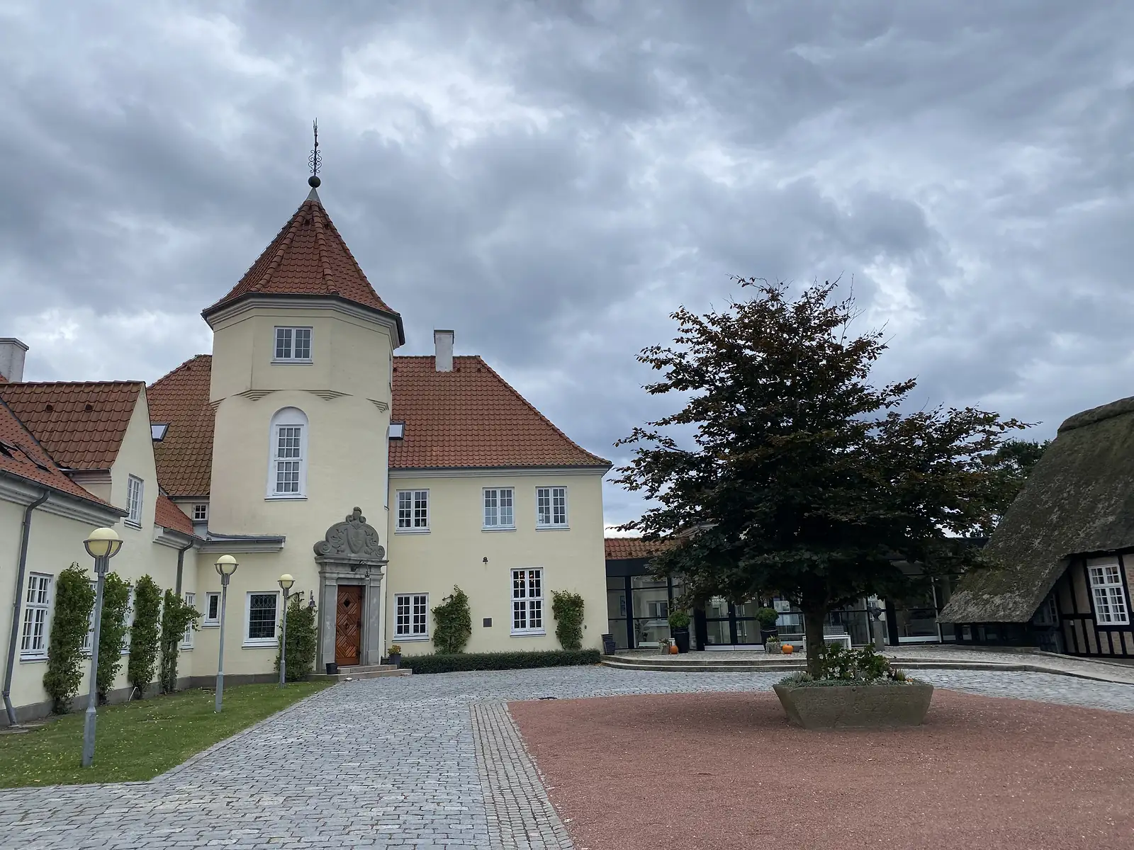 A cream-rendered manor with an octagonal tower and red-tiled roofs above a cobbled courtyard with a copper beech in a stone planter.
