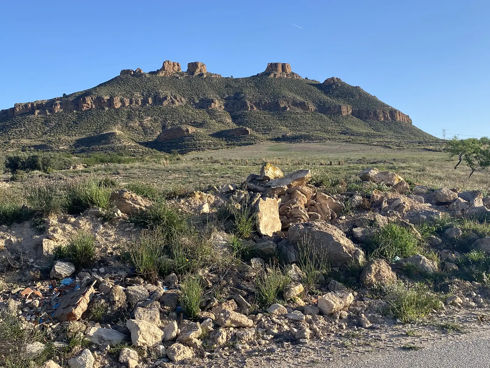 A flat-topped hill crowned with eroded rock towers, seen across scrub and a roadside bank of dumped rubble.