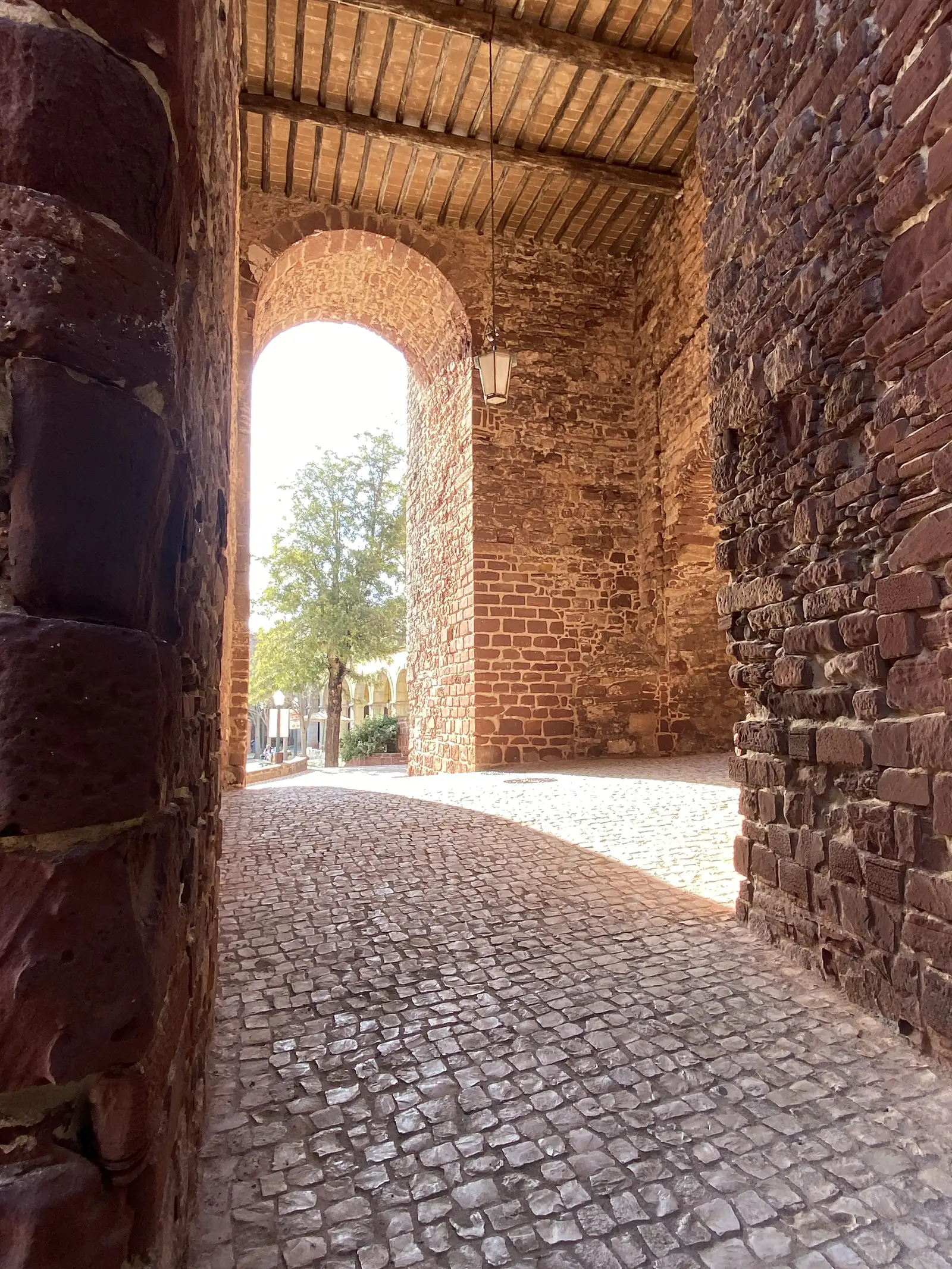 Cobbled passageway running through the red sandstone gatehouse of a castle, a timber-and-brick ceiling overhead and a tree lit by sunlight beyond the far arch.