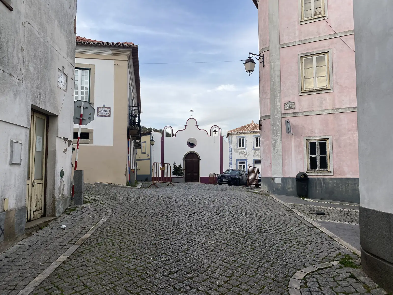Cobbled square between a pink-washed corner house and older whitewashed buildings, closed off at the end by a small white chapel with a scalloped baroque gable and a dark arched door.