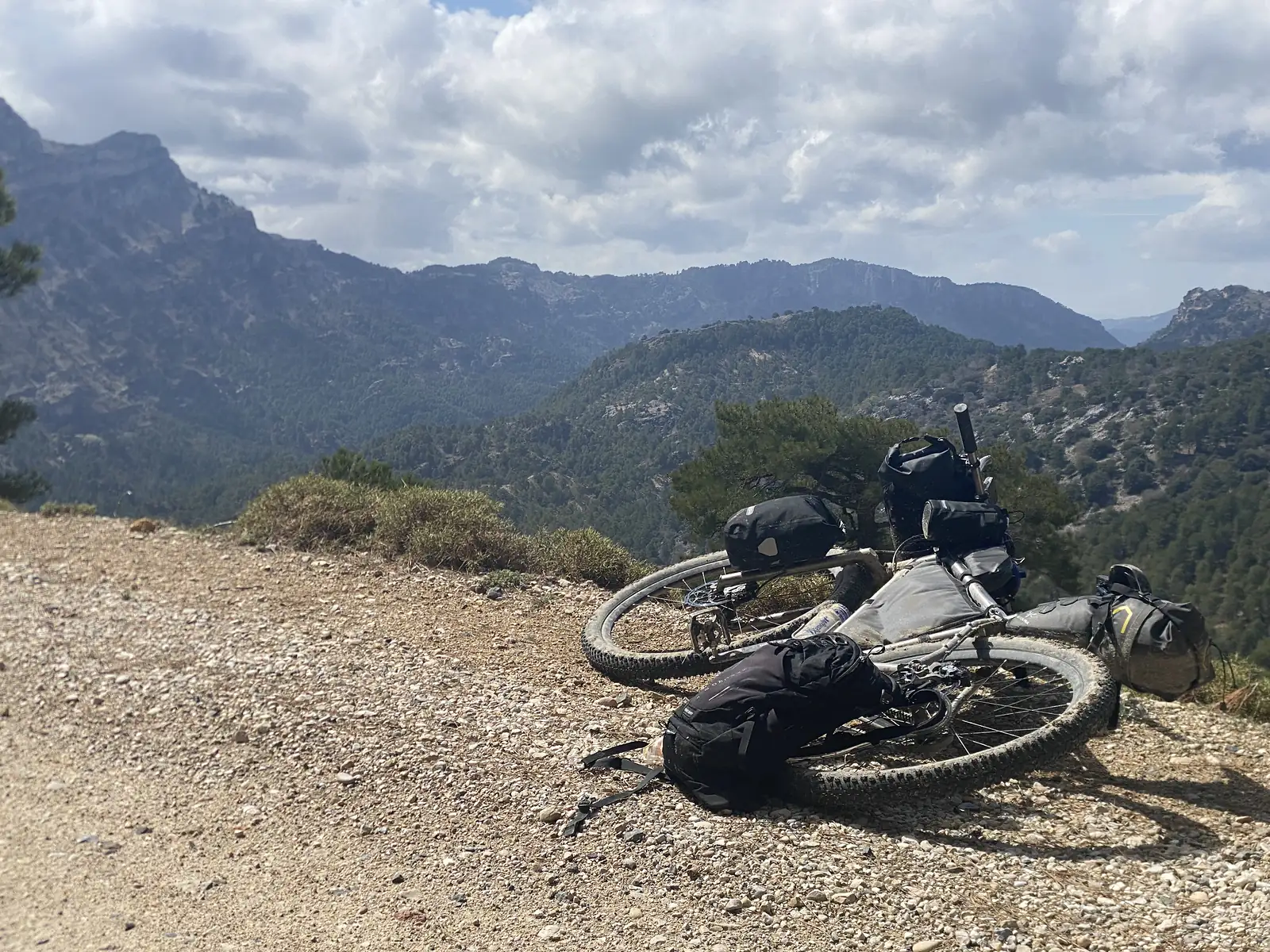 A loaded bikepacking bike laid on its side on a gravel track at a mountain viewpoint, with pine-covered ridges falling away beyond.