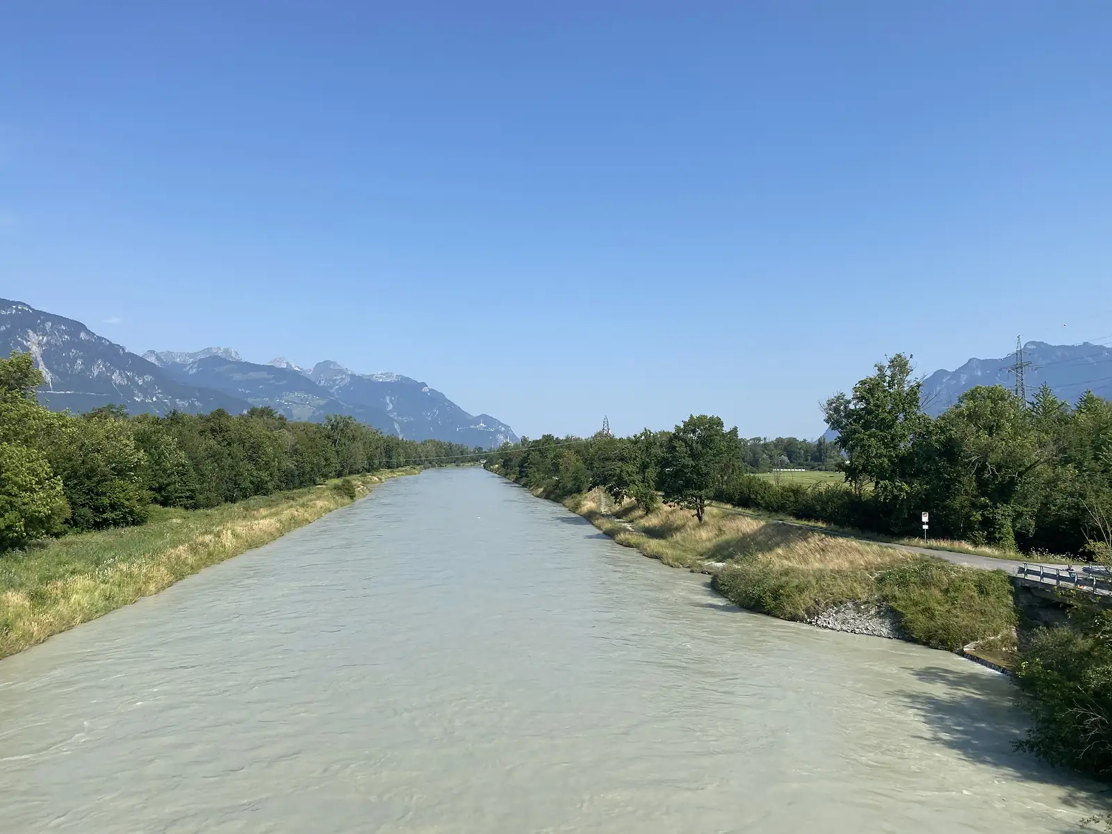 A straight, fast-flowing glacial river channel bordered by trees and a cycle path runs towards distant mountains under a clear sky.