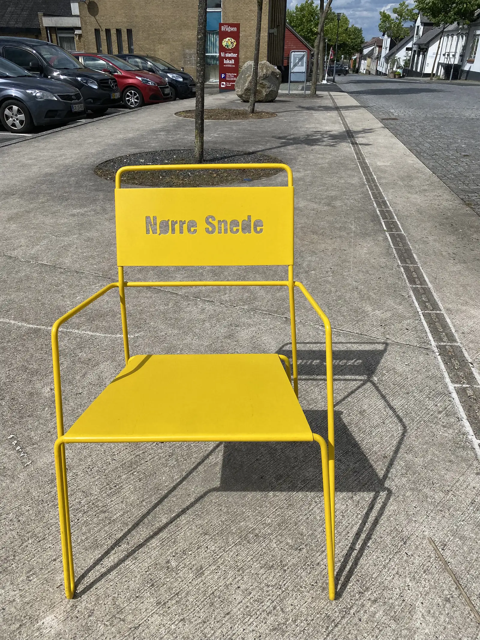 A large yellow steel chair on a town pavement with "Nørre Snede" cut through its backrest, the name thrown as a shadow on the ground.