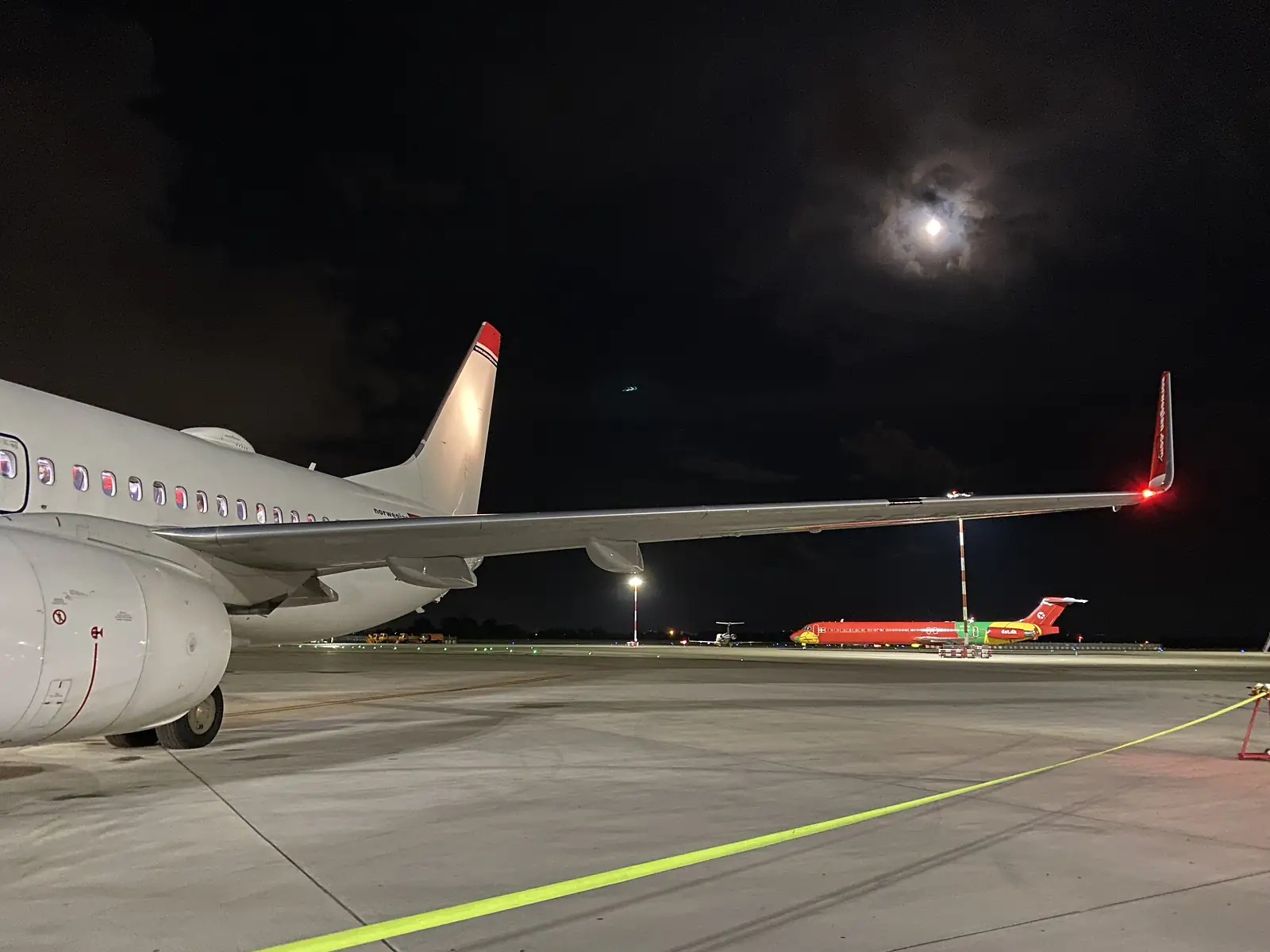 A moonlit view along an aircraft wing on the tarmac at night, with a red-liveried plane taxiing in the distance.