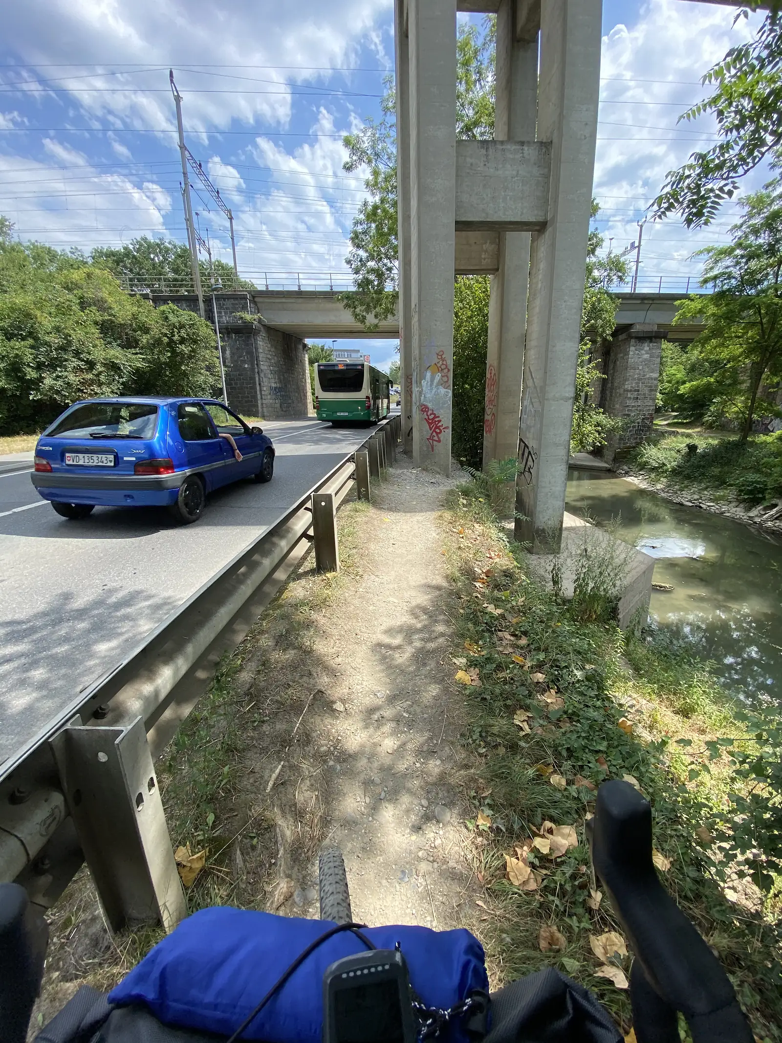 A path passing beneath a road and rail viaduct alongside a stream, with a bus and a small blue car crossing the bridge above.