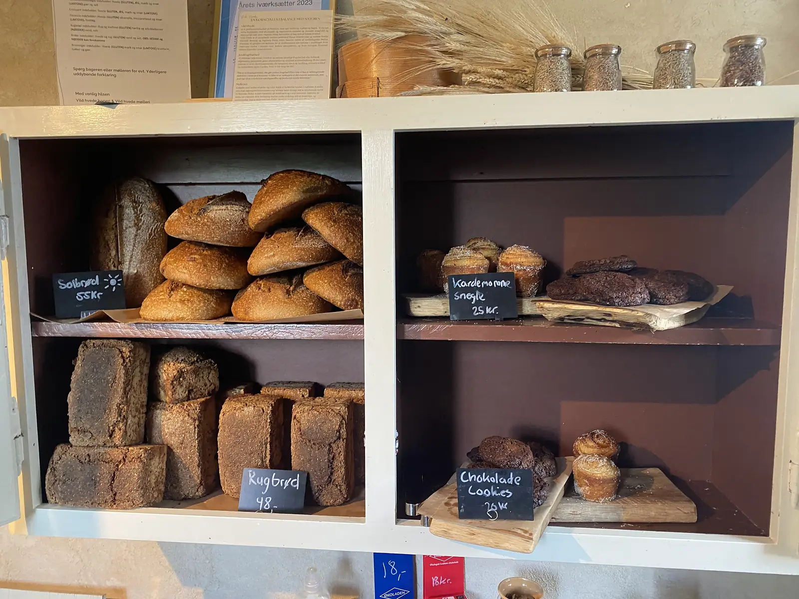 A bakery cabinet of sourdough loaves and dark rye bread with chalked price tags, and cardamom buns and chocolate cookies on wooden boards.