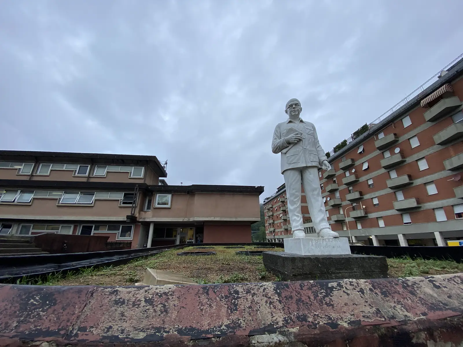 A white statue of a bespectacled man in jacket and trousers, holding a book, stands on a plinth in a paved courtyard between apartment blocks.