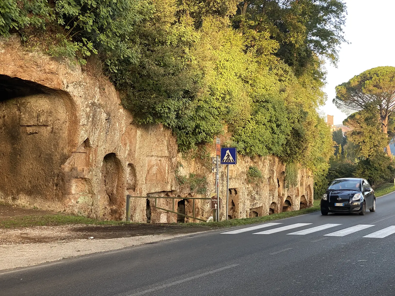 A roadside cliff of soft rock riddled with rows of carved niches, likely ancient tombs, with a car passing a pedestrian crossing and a brown sign reading "Anfiteatro mt 35".