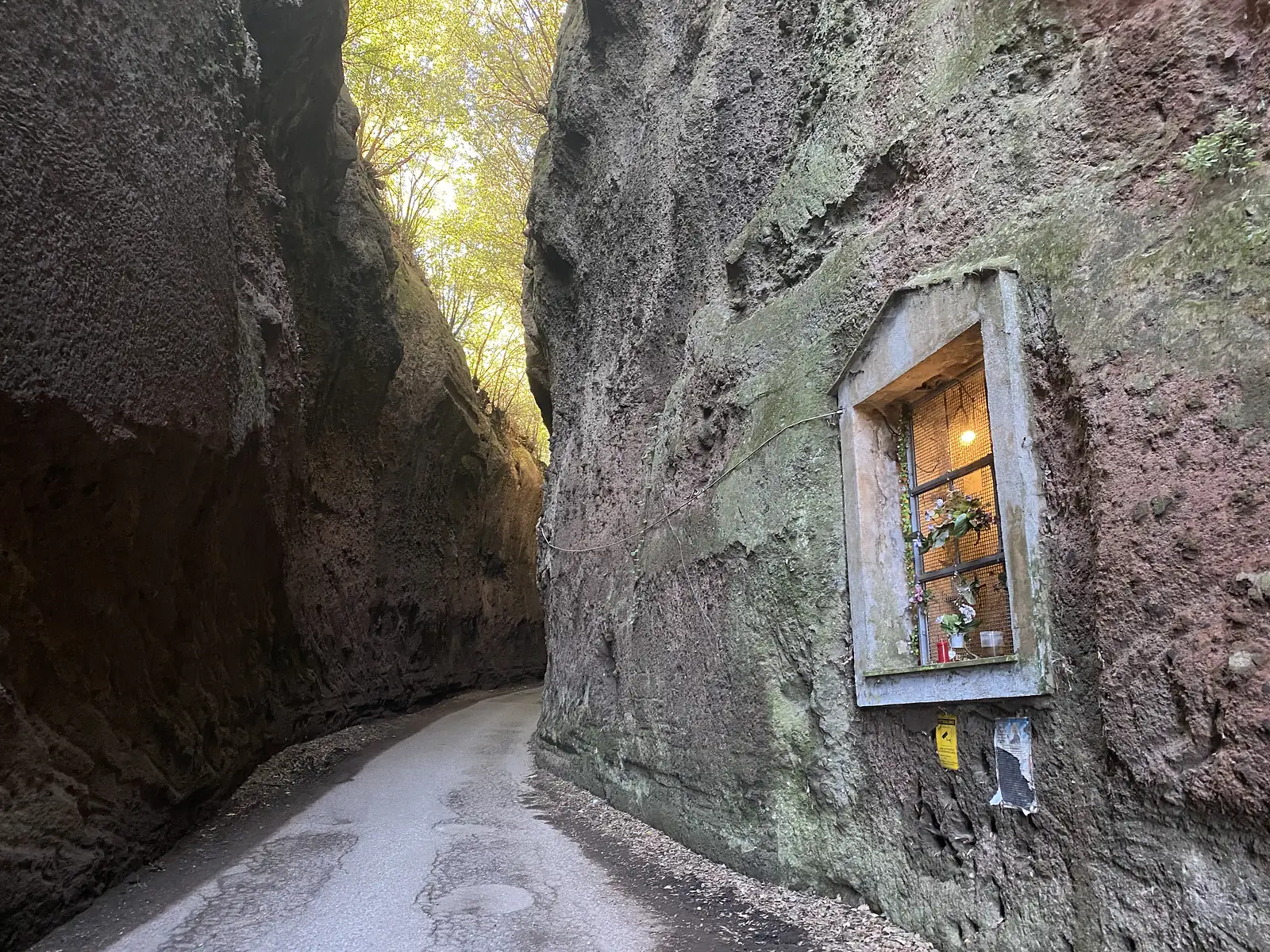 A small illuminated wayside shrine with candles and flowers set into a niche in the rock wall of a deep sunken lane cut through tufa.
