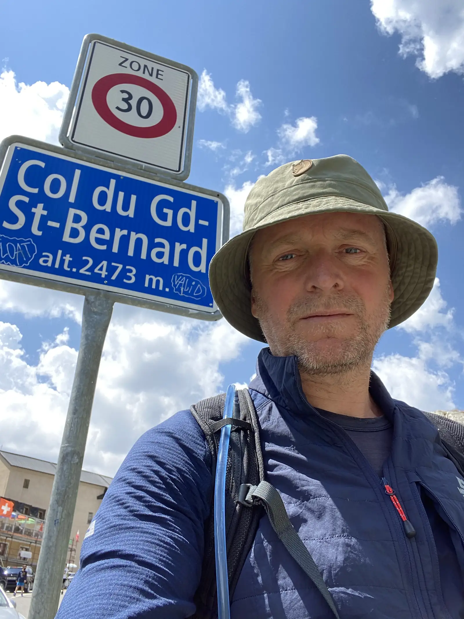 
A hiker in a bucket hat taking a selfie beside a blue sign reading "Col du Gd-St-Bernard alt.2473 m." and a Zone 30 speed-limit sign.
