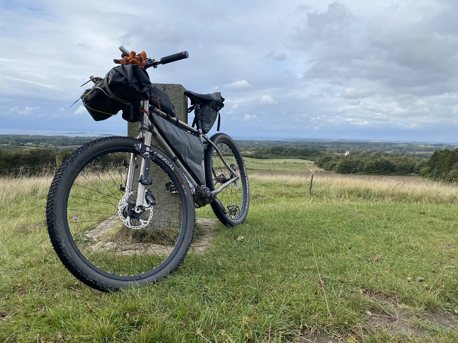 A loaded bikepacking bike leaning against a stone marker on a grassy hilltop, with fields, woods and a distant sea horizon.