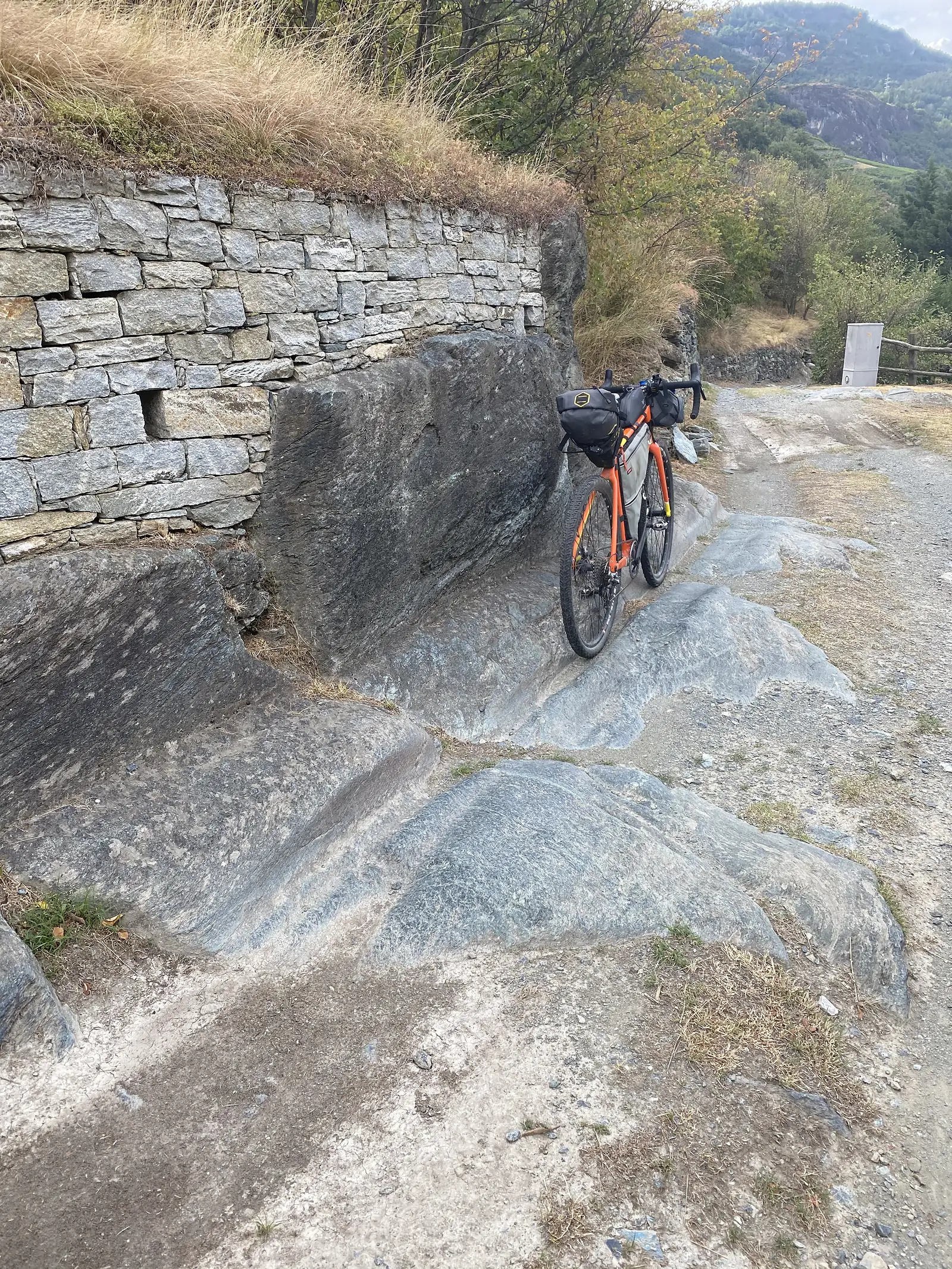 A loaded orange gravel bike propped against a low dry-stone wall on a rocky outcrop beside a gravel track.