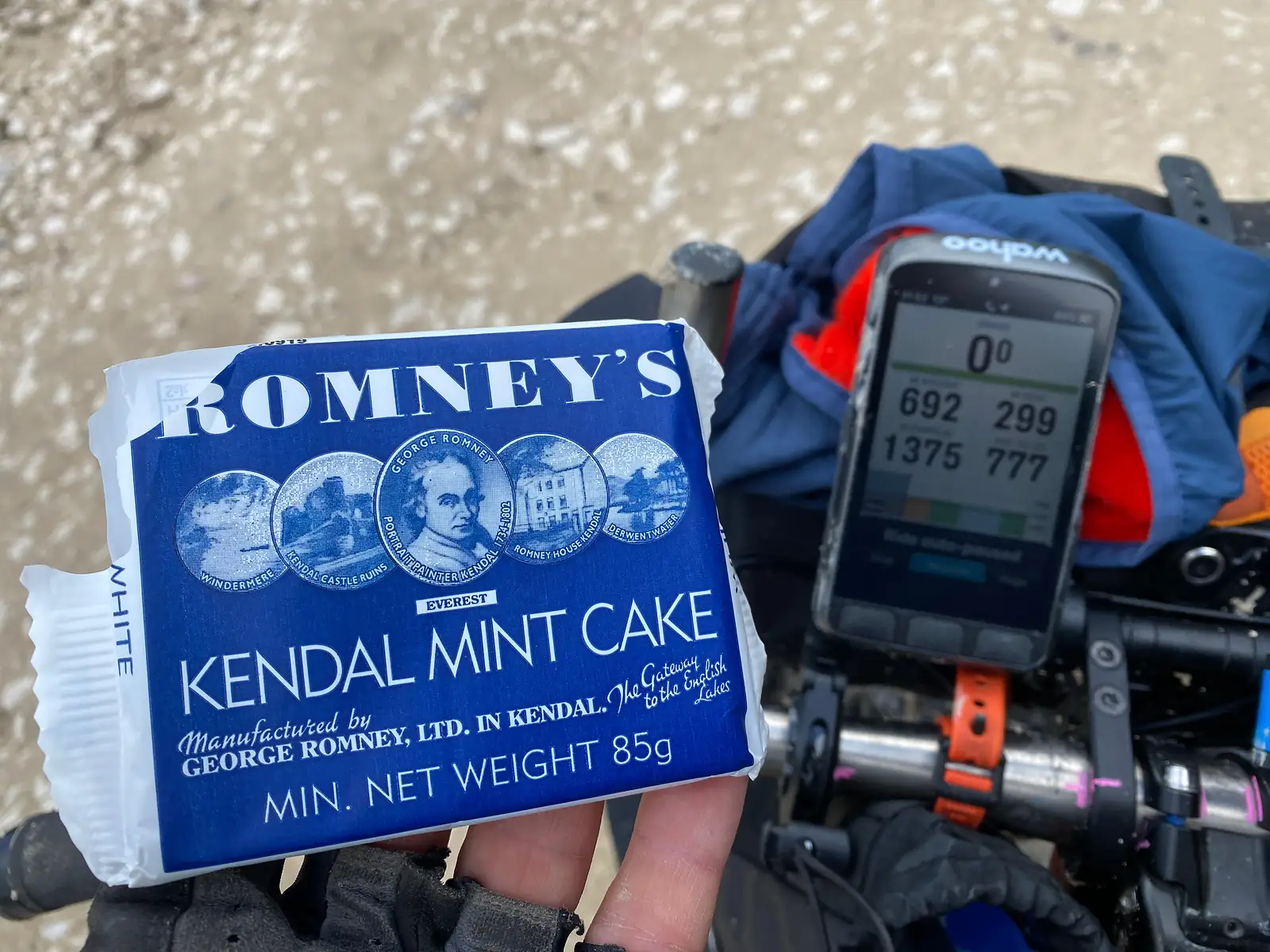 A gloved hand holding a bar of Kendal mint cake above the handlebars, beside a bike computer showing the day's numbers.