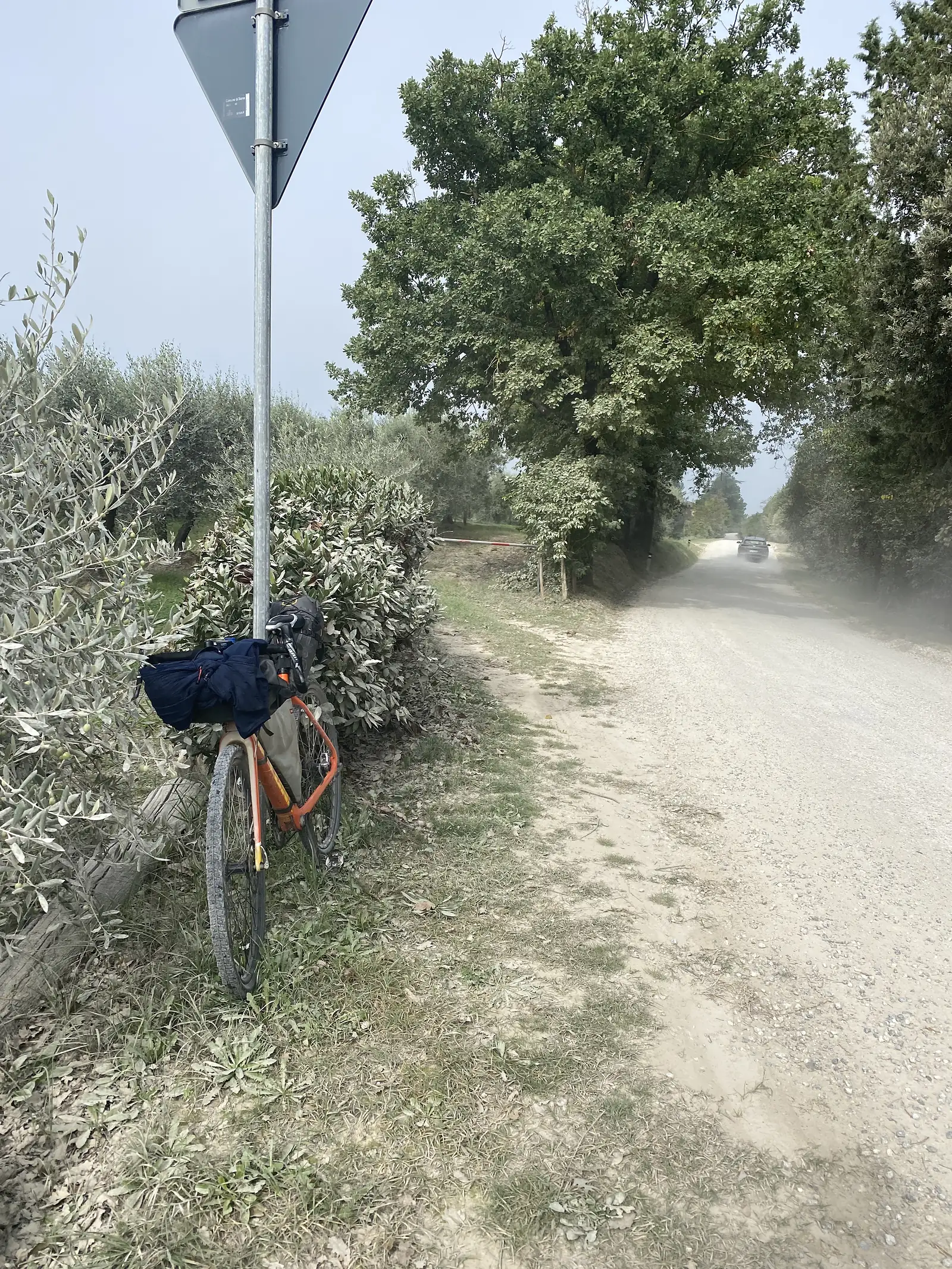 A loaded orange bikepacking bike leaning against roadside shrubs beside a signpost, with a gravel track and a figure walking in the distance.