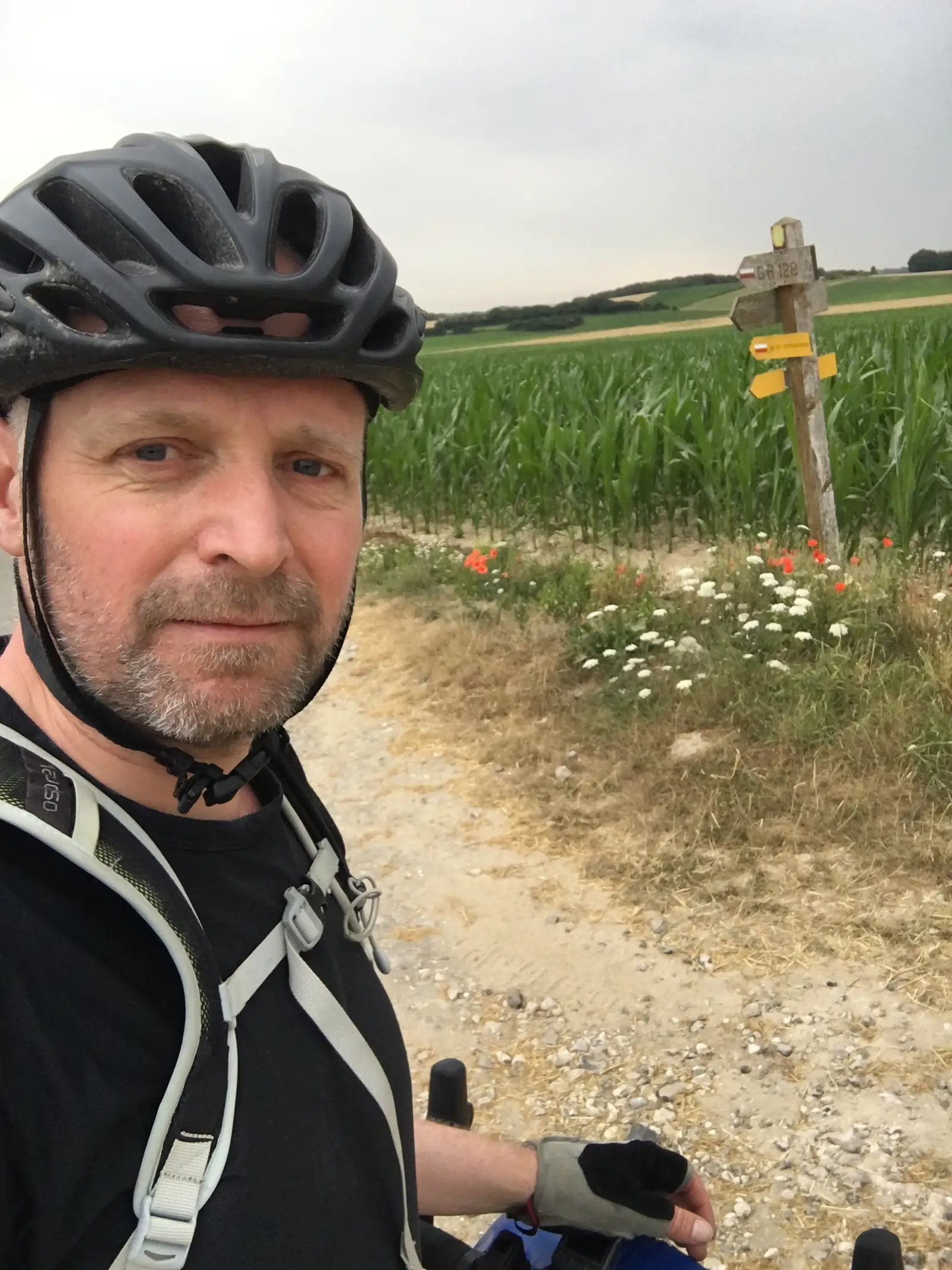 A cyclist in a helmet and fingerless gloves takes a selfie beside a wooden waymarker post reading 'GR 128', its yellow and red-and-white markers pointing along a gravel track past poppies and wheat.