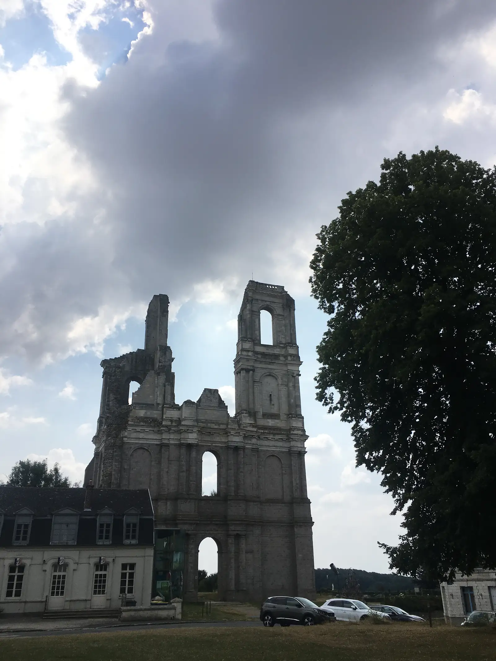 The ruined twin towers of a gutted stone church standing roofless against a cloudy sky, with parked cars and a house visible below.