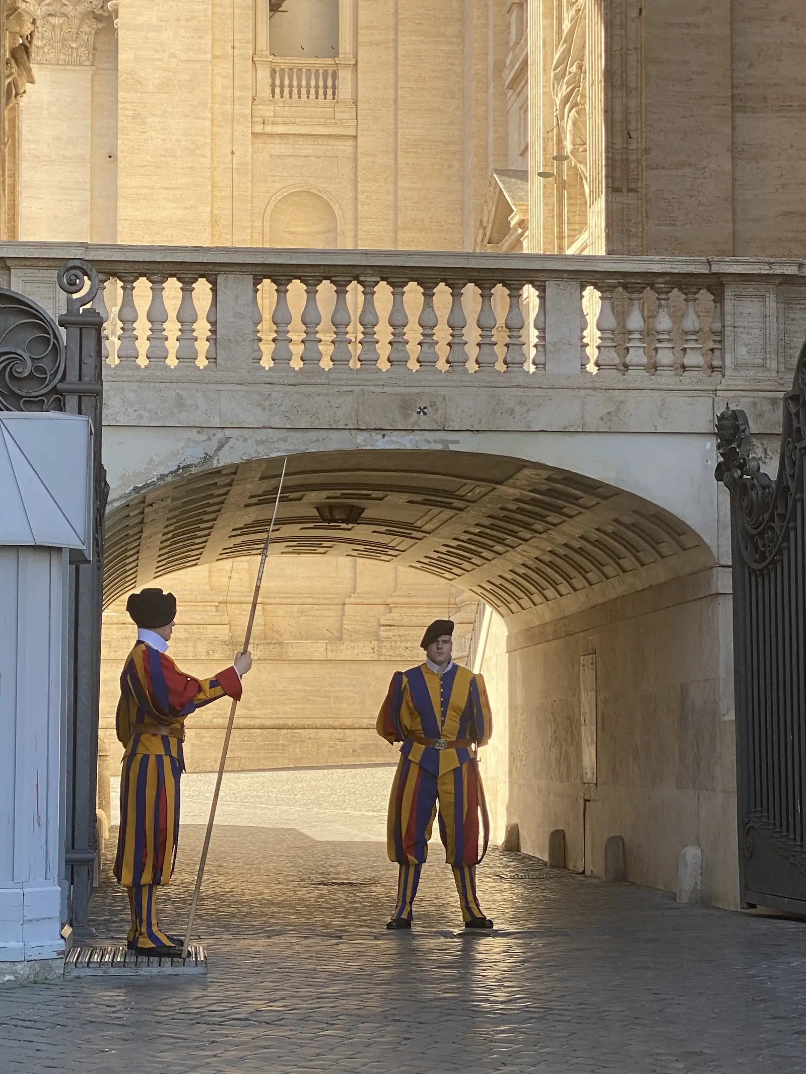 Two Swiss Guards in blue, red and yellow striped uniform stand watch with a halberd beneath a stone archway at the Vatican.