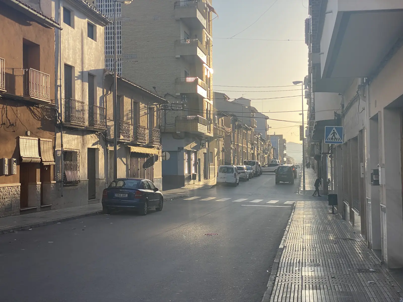 A wide town street at sunrise, low sun flaring between apartment blocks and older balconied houses, with a few parked cars and a lone walker.