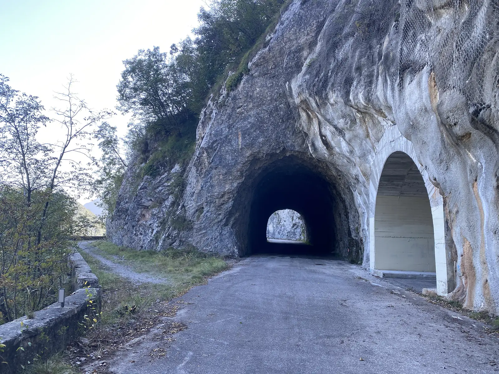 An unlit tunnel bored through rock, beside an open concrete avalanche gallery, on a quiet single-track mountain road.