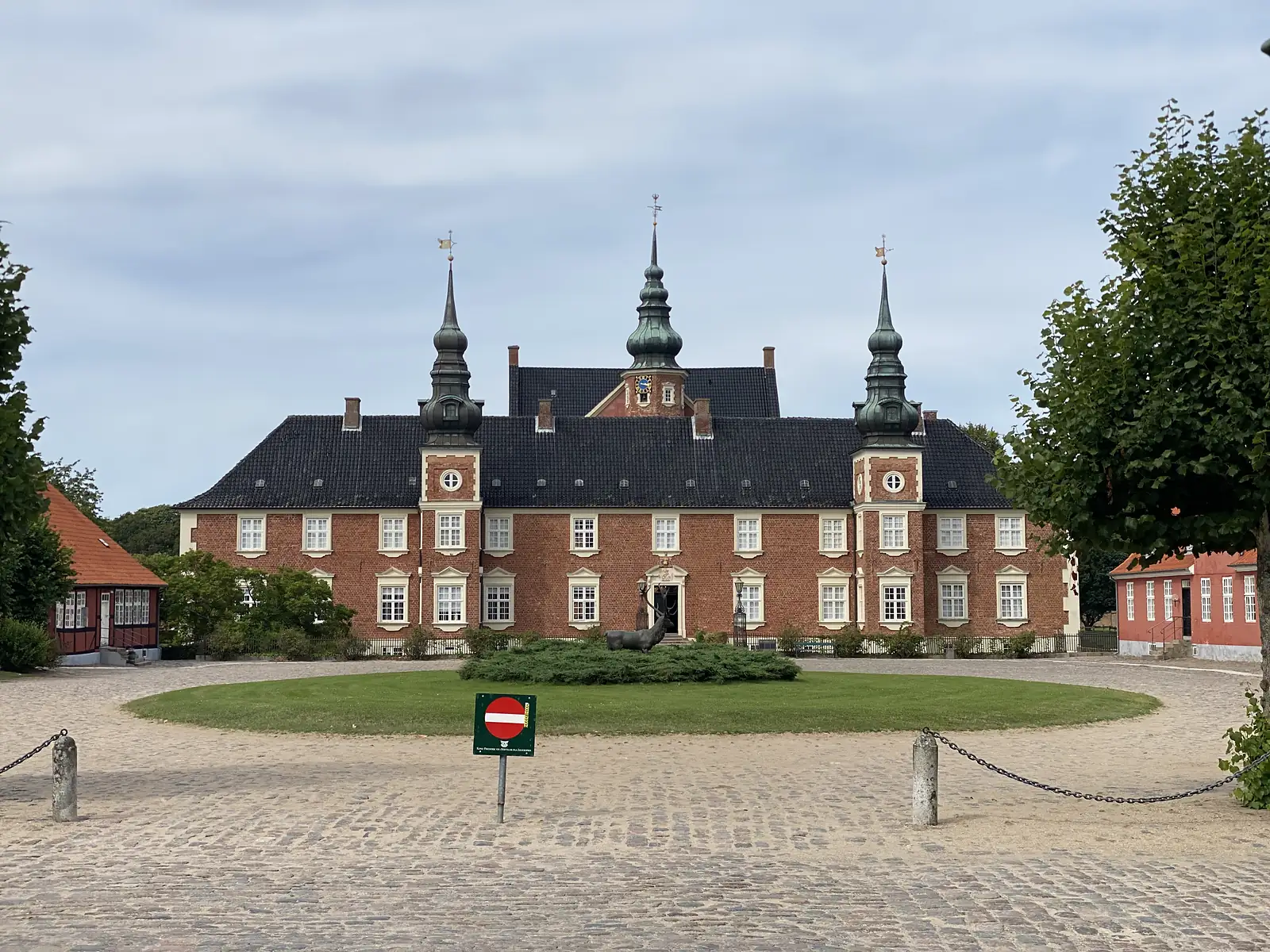 Jægerspris Slot, a red-brick manor house with three copper-green spires, seen across its cobbled forecourt and circular lawn.