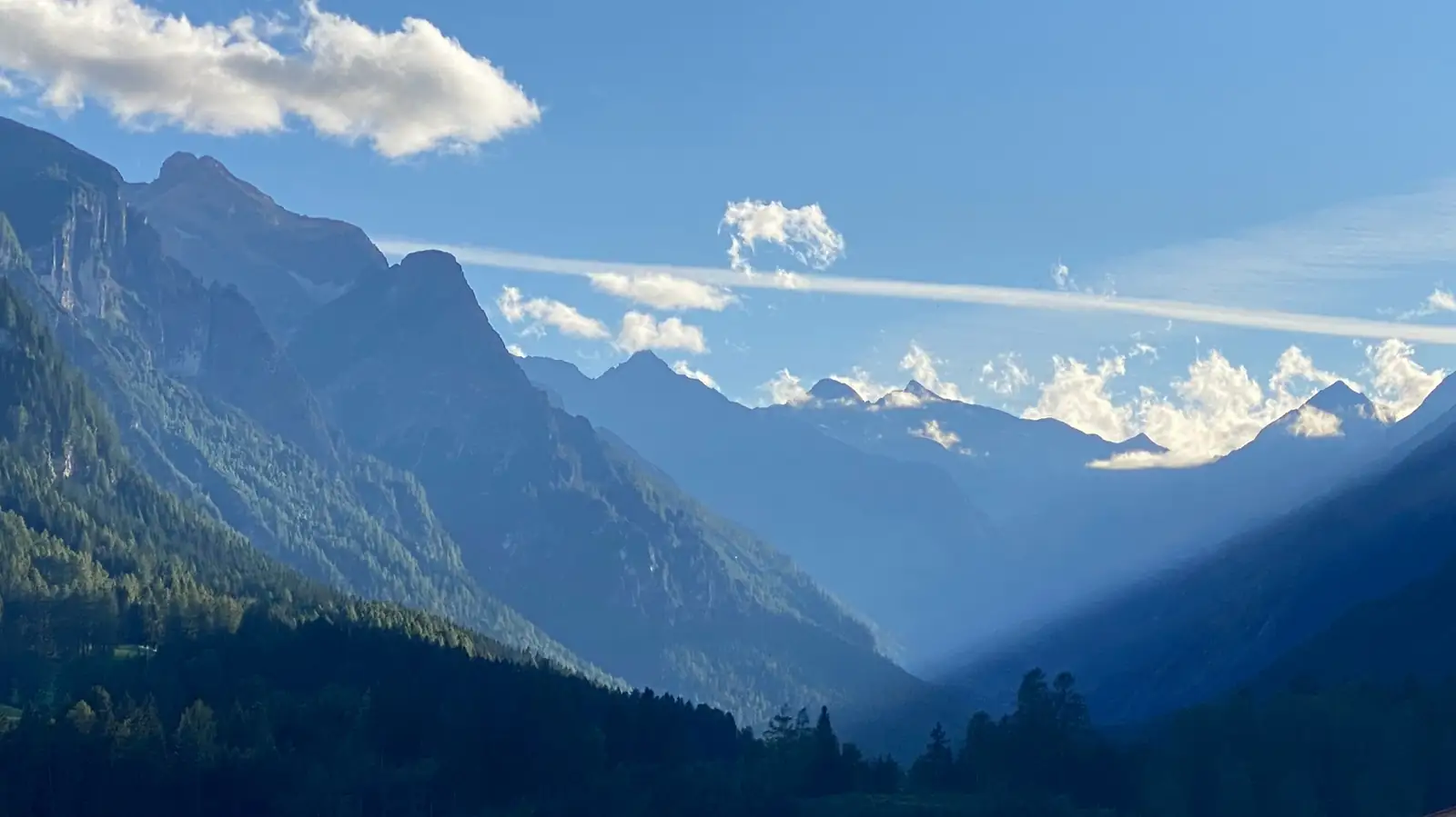 Layered mountain ridges recede into haze above dark forest, with low cloud caught along the skyline.