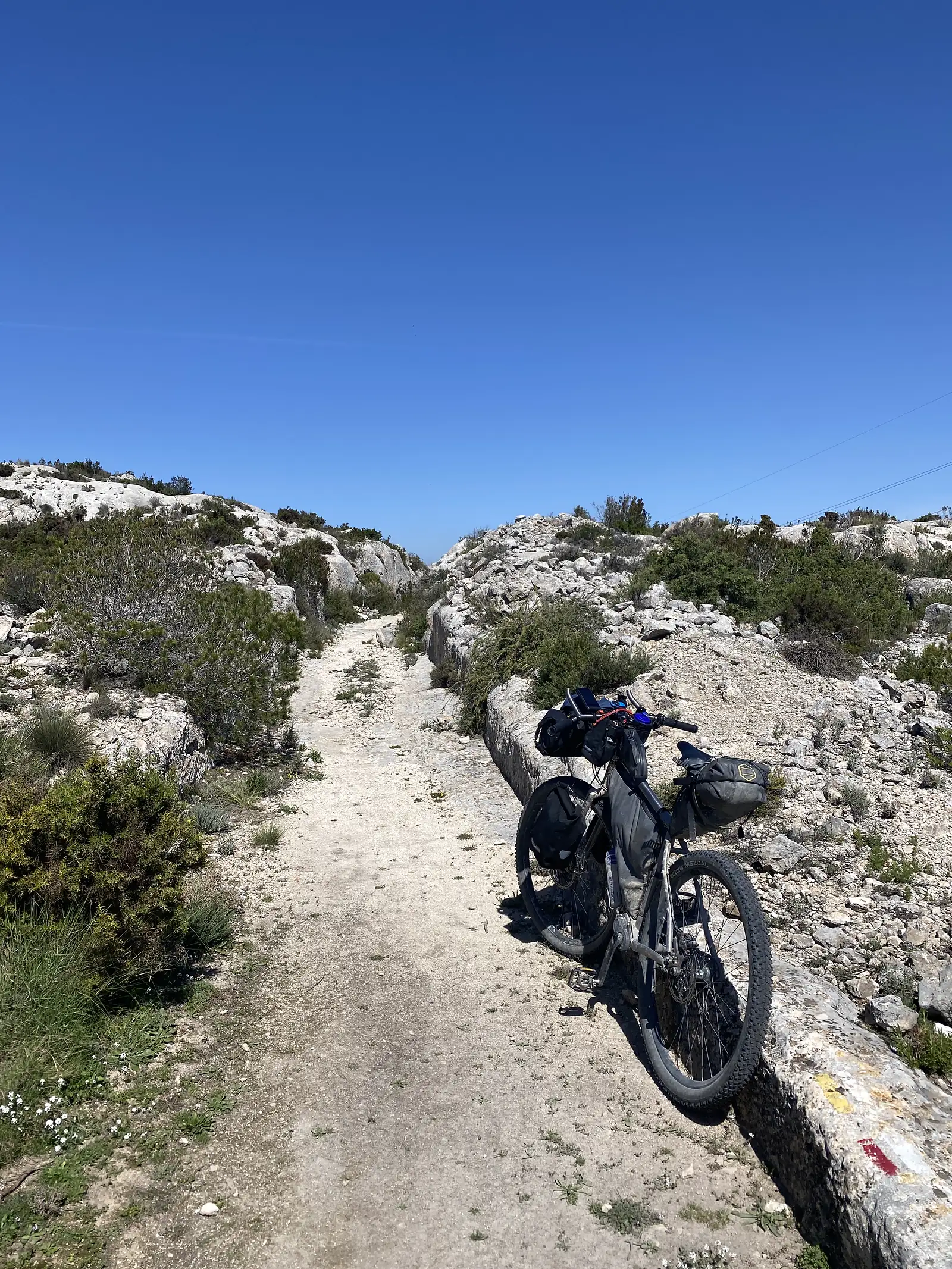 A loaded bikepacking bike leaning against a low wall beside a rough limestone track, a red-and-white waymark painted on the stone.