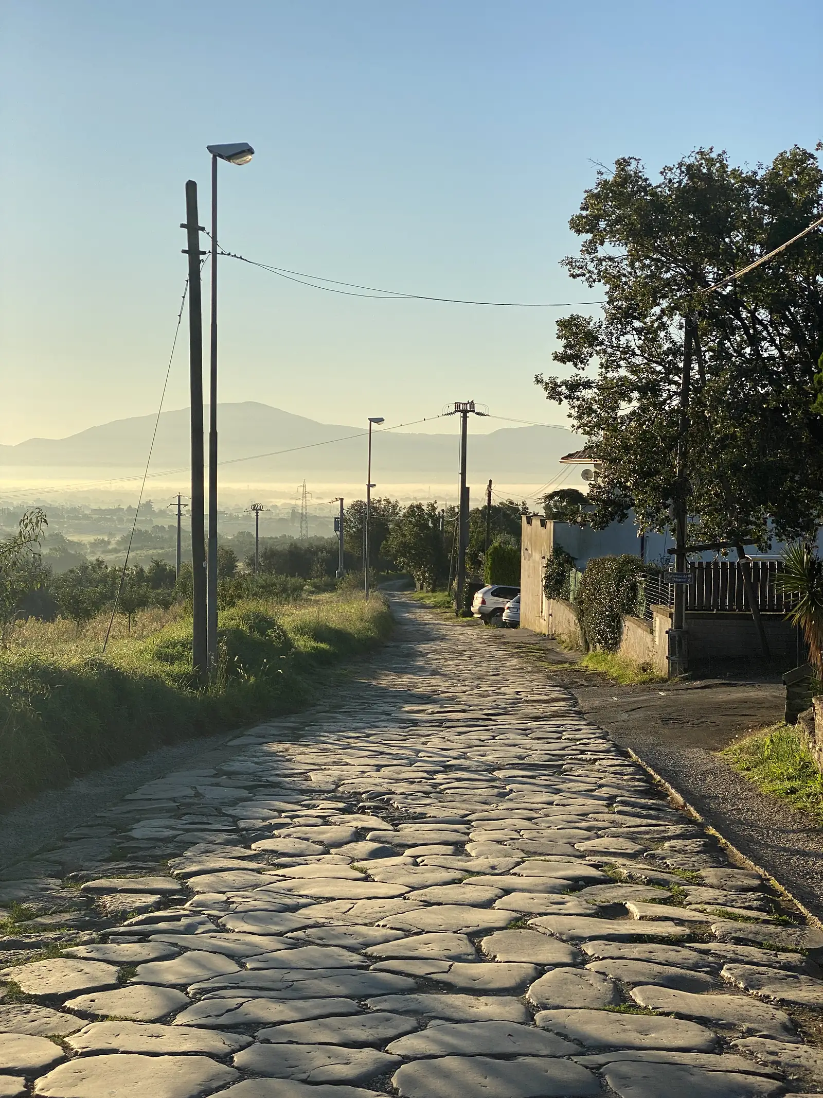 Cobbled lane past a gated house with a parked car, a line of telegraph poles receding towards hazy hills.
