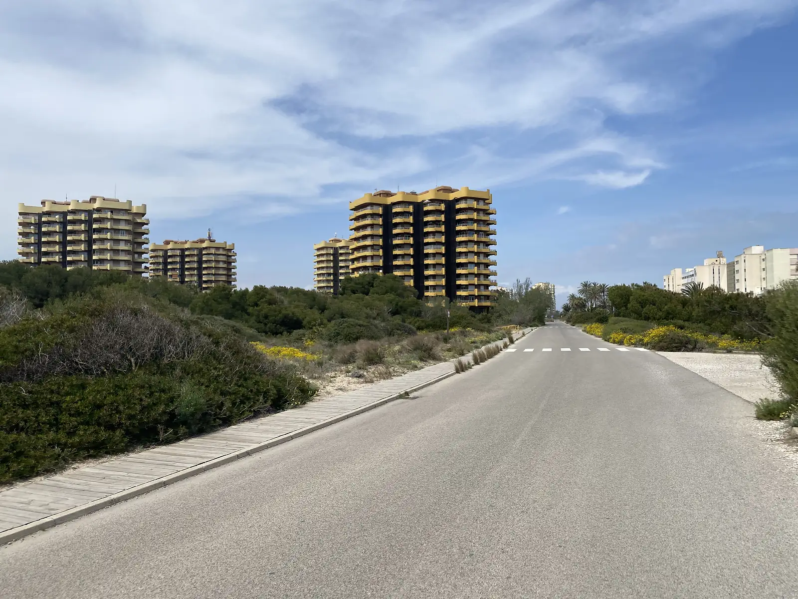 An empty seafront road with a boardwalk alongside running past dune scrub towards yellow high-rise apartment blocks.